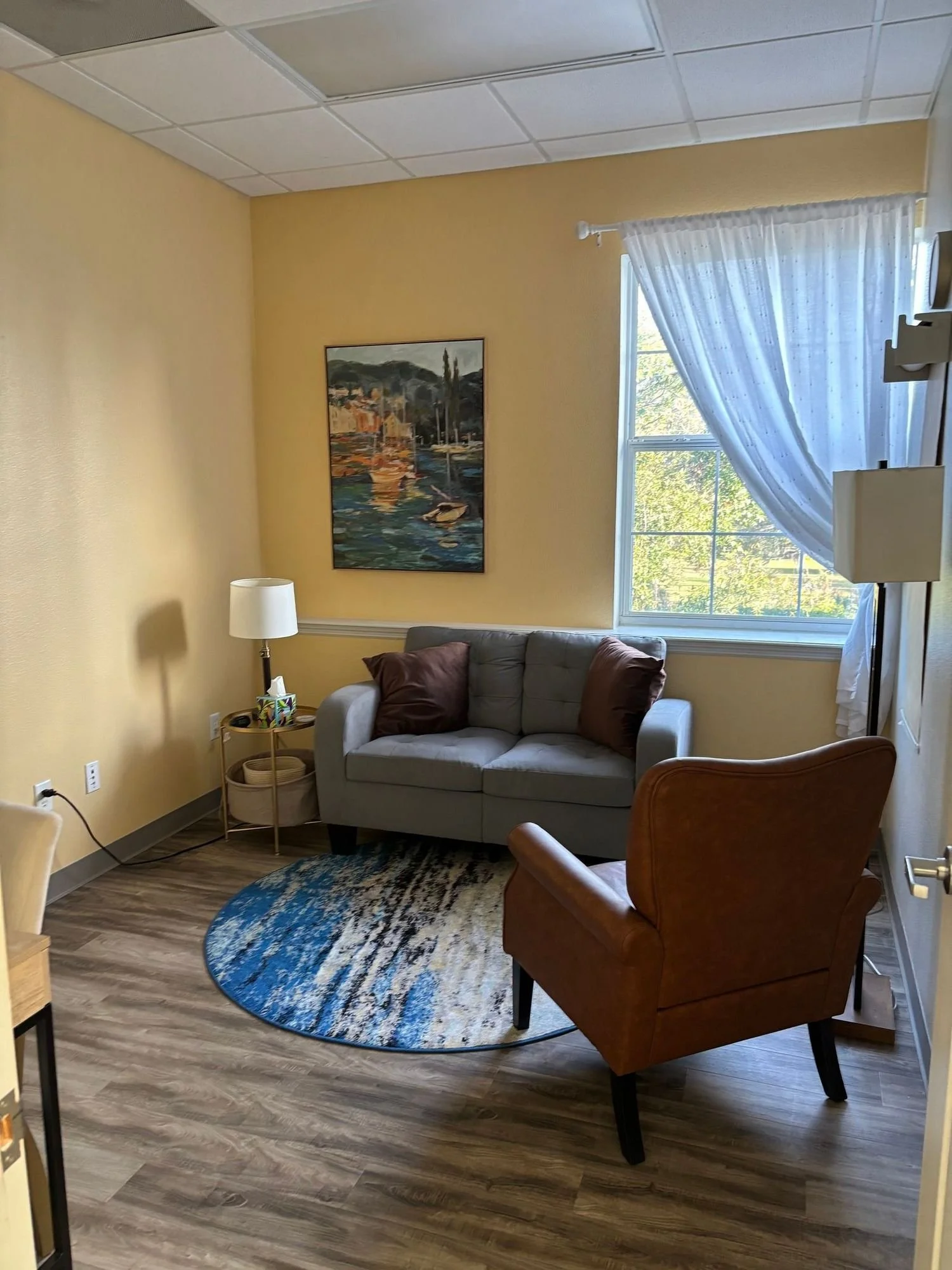 A cozy living room with a small, light gray sofa adorned with two brown pillows. A round, blue patterned rug lies beneath the sofa. To the side, there is a floor lamp with a white shade next to a small table holding tissue and other items. The wall behind the sofa is painted a warm yellow and features a framed painting of a harbor scene with boats and trees. To the right, a window with sheer white curtains is letting in natural light, revealing trees outside. A wood floor completes the room.