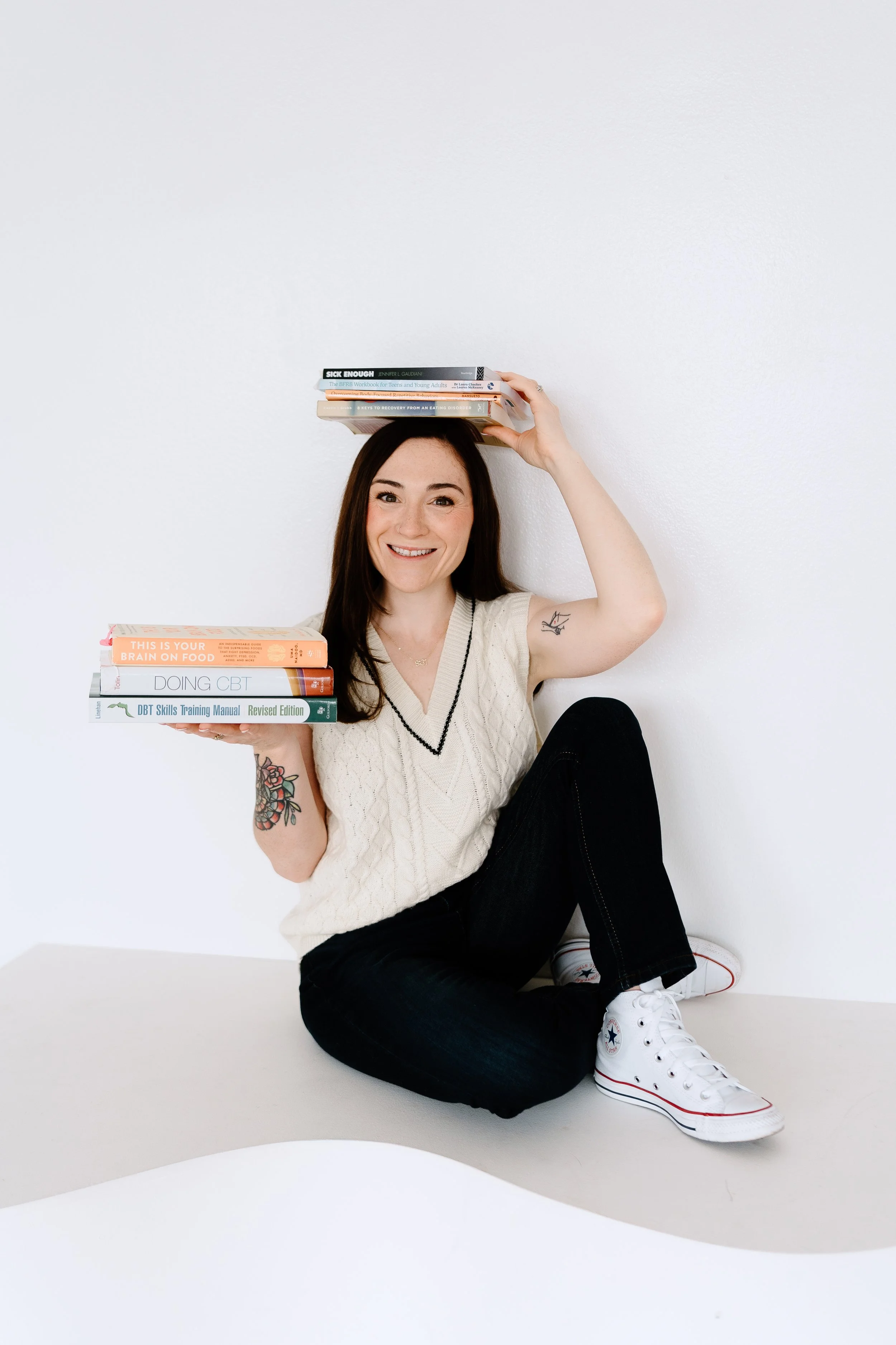 A smiling woman with long dark hair, tattoos on her arm, wearing a beige sweater, black jeans, and white Converse sneakers, sitting on the floor against a plain white wall. She is holding a stack of books in one hand and balancing another stack of books on her head.