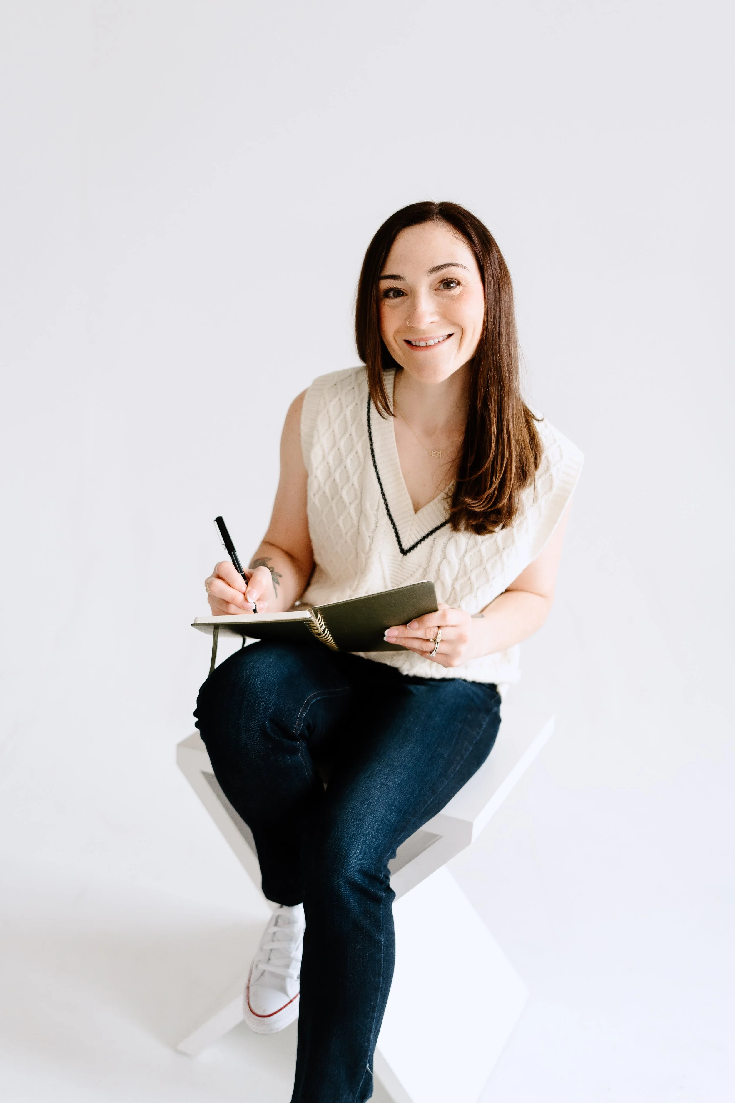 A young woman with long brown hair, smiling, wearing a cream sleeveless sweater and dark jeans, sitting on a white block, holding a pen and a black notebook, against a plain white background.