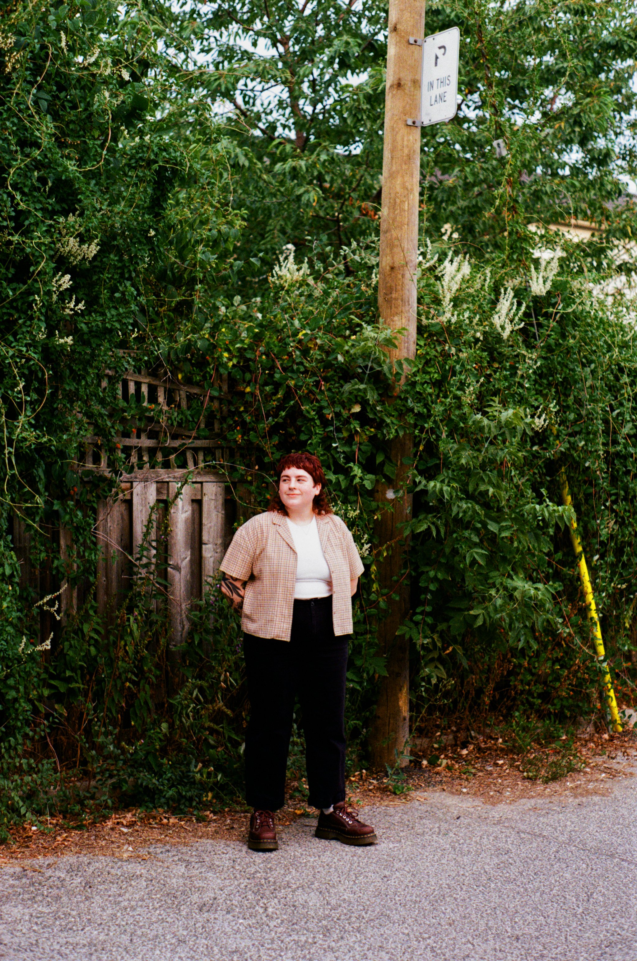 Kristi pictured with red hair, wearing a beige plaid shirt over a white top, black pants, and dark loafers, standing on a sidewalk next to a wooden fence and dense green foliage.