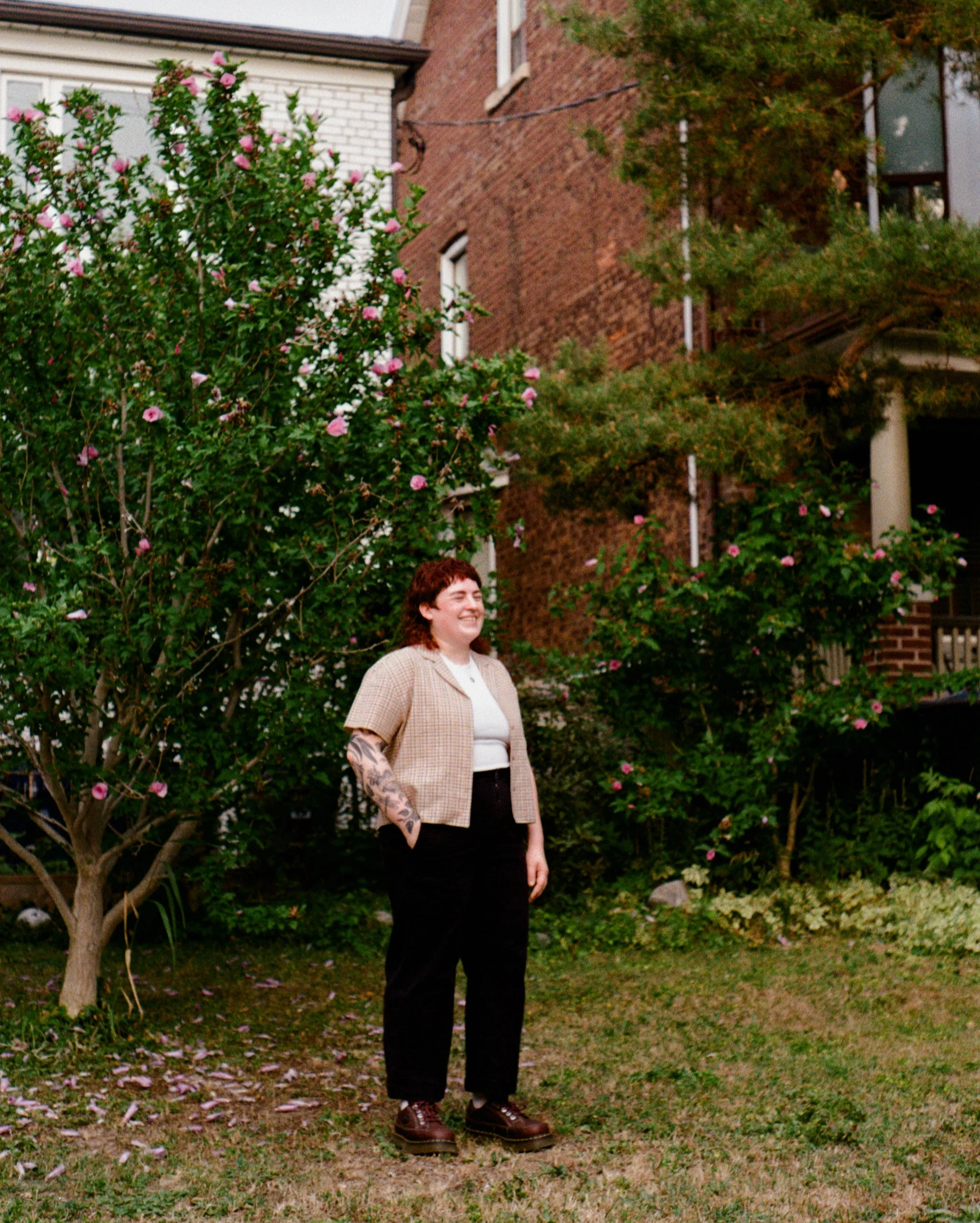 Kristi pictured with red hair and tattoos on their right arm standing in front of a blooming pink-flowered tree, smiling, in a yard near brick buildings.