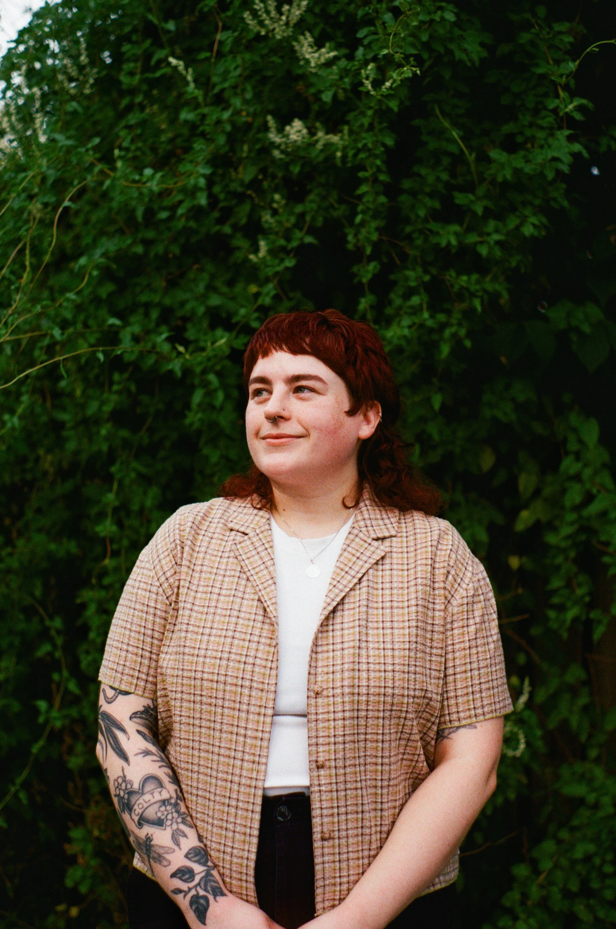 Kristi pictured with red hair, a septum piercing, and tattoos on left arm, standing outdoors in front of green foliage. They are wearing a plaid short-sleeve shirt over a white top and looks to the side with a slight smile.