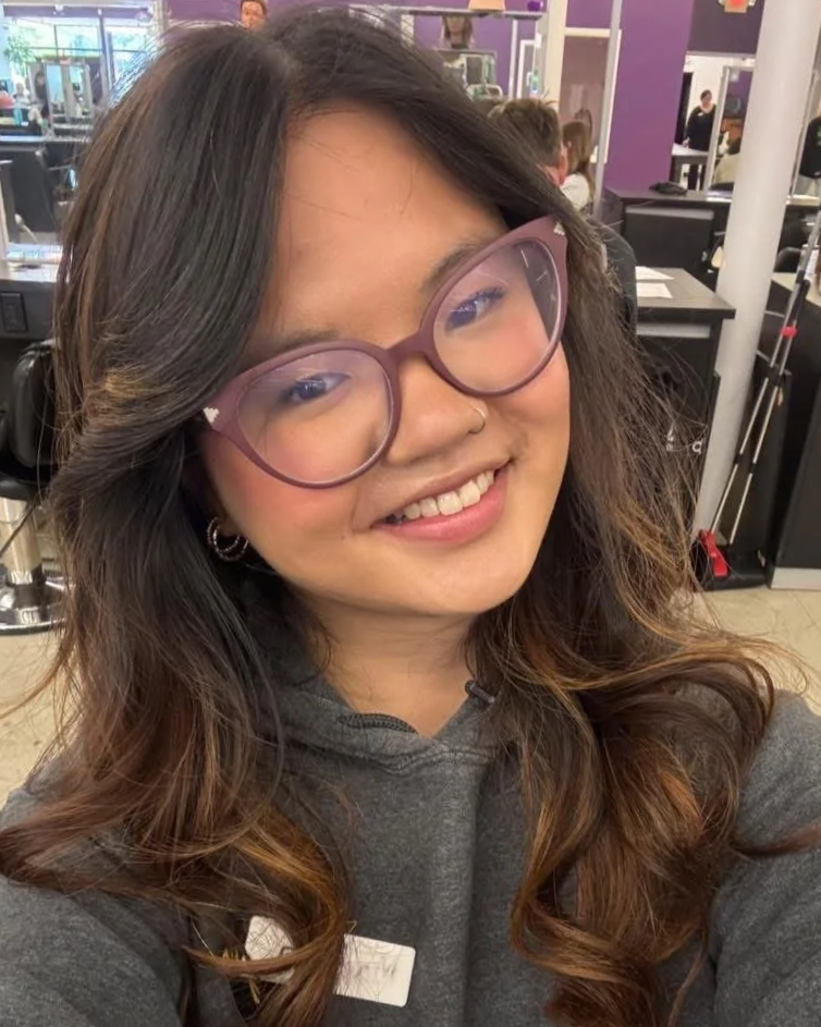 A young woman with curly brown hair, wearing pink glasses, a gray hoodie, and earrings, smiling in a gym.