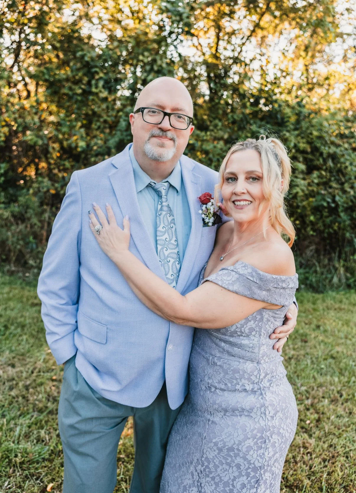 A couple dressed in formal attire, standing outdoors in front of green foliage, embracing each other and smiling at the camera.