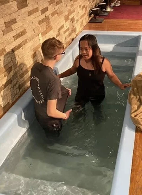 A woman and a young boy are in a baptismal pool inside a church, with the woman appearing to have been baptized and the boy holding her hand.