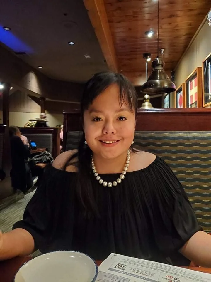 Woman with long dark hair and a pearl necklace smiling at the camera in a restaurant with wood-paneled ceiling and framed pictures on the wall.