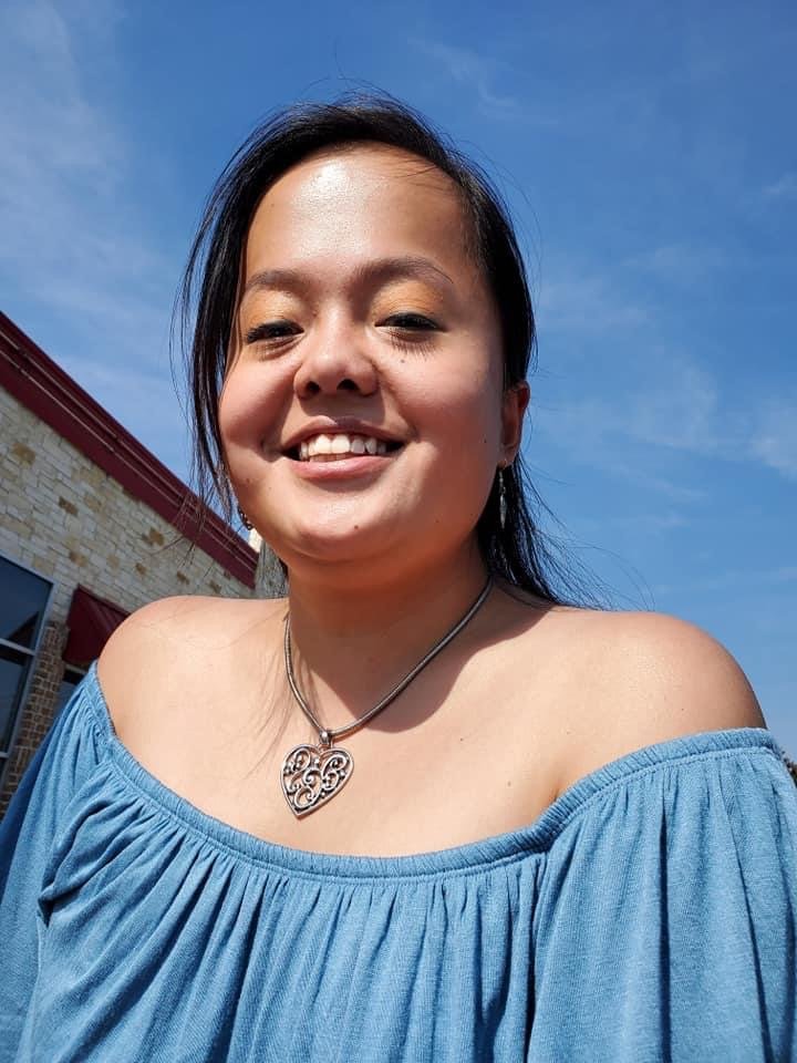 A woman smiling outdoors against a blue sky, wearing a blue off-the-shoulder top and a necklace with a heart-shaped pendant.