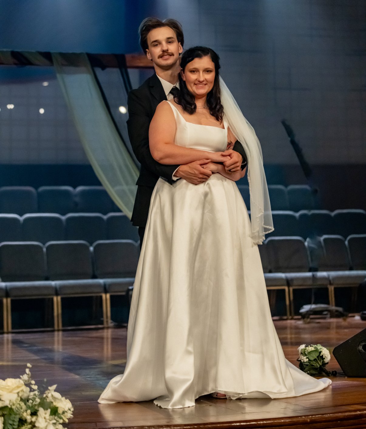 A bride and groom standing together on a stage in a theater, with seating in the background. The bride is wearing a white wedding gown and veil, and the groom is in a black suit, white shirt, and black tie. They are embracing each other, smiling at the camera.