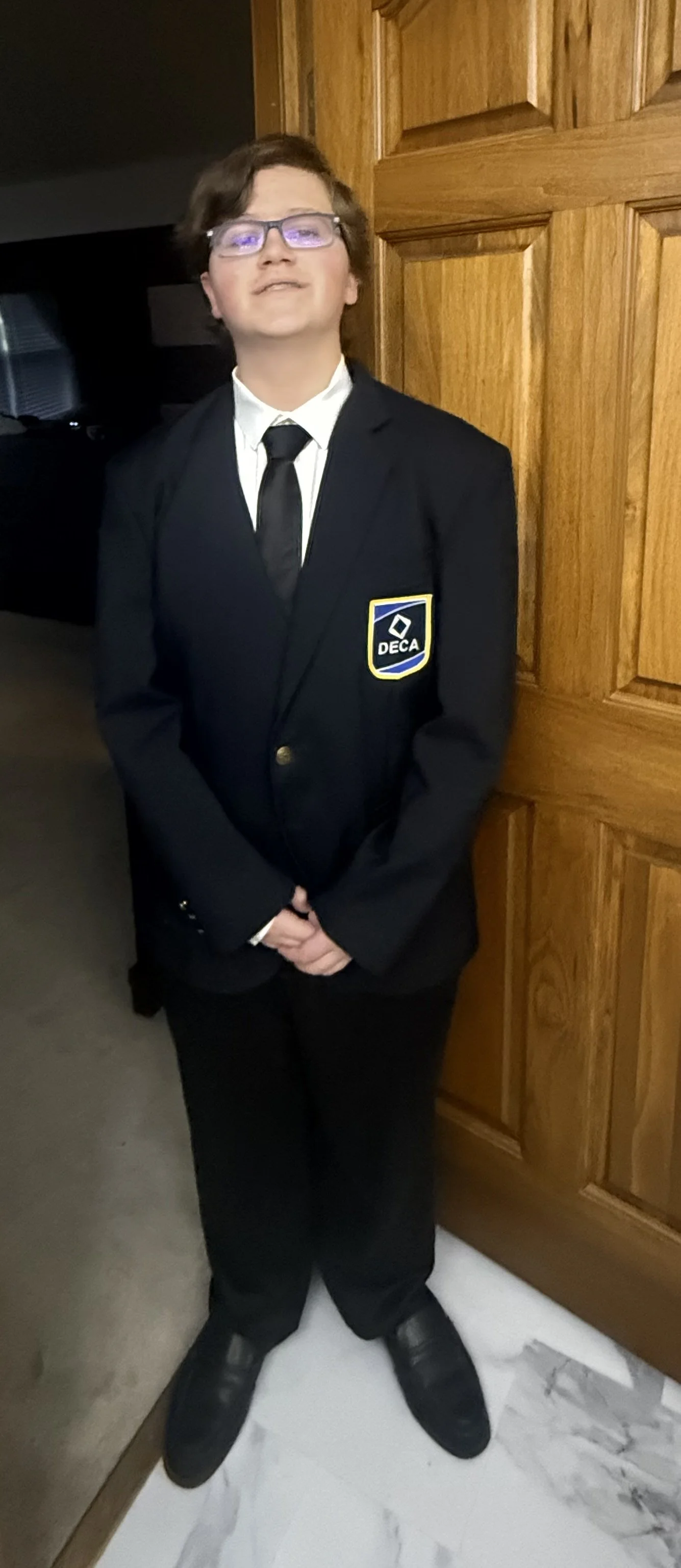 A young boy dressed in a black suit with a DECA badge, white shirt, black tie, glasses, standing indoors next to wooden wall panels.