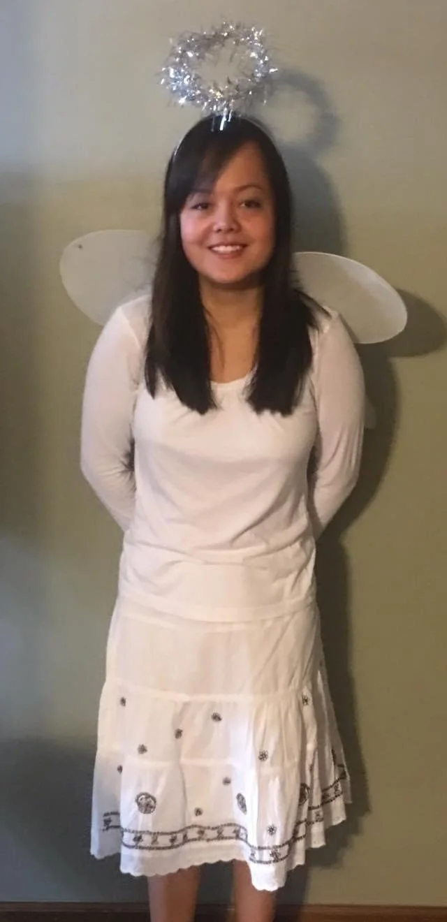 Woman dressed as a fairy, with white fairy wings, a white dress, and a silver tinsel halo on her head, standing against a plain wall.