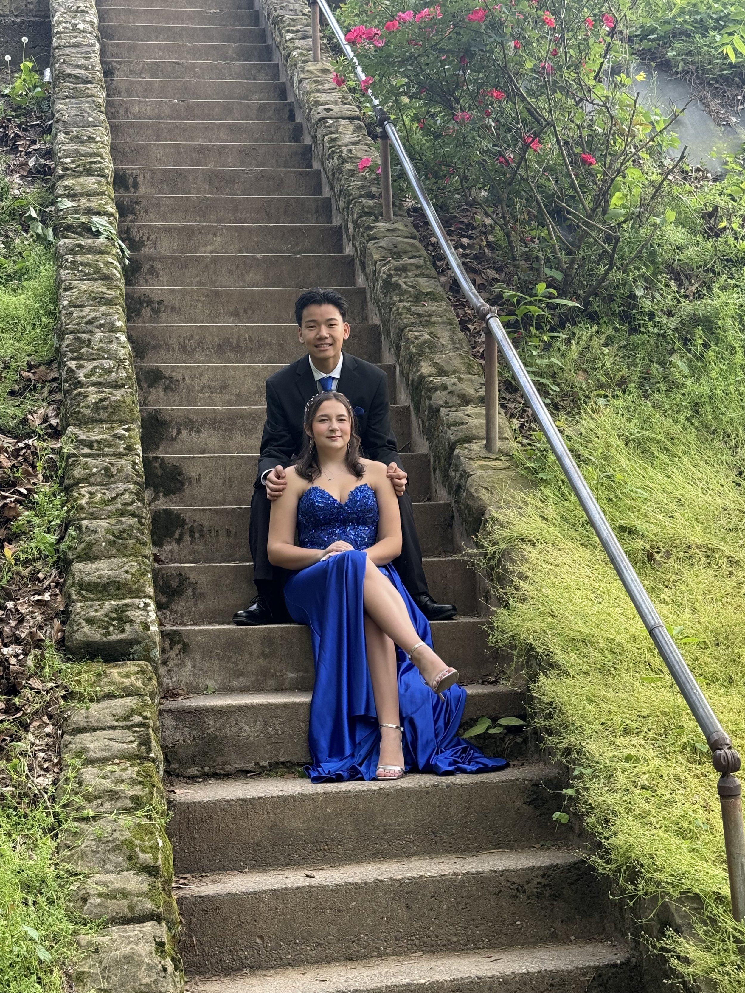 A young man and woman sitting on outdoor stone staircase, surrounded by greenery and pink flowers. The woman is in a blue evening gown, and the man is in a black suit.