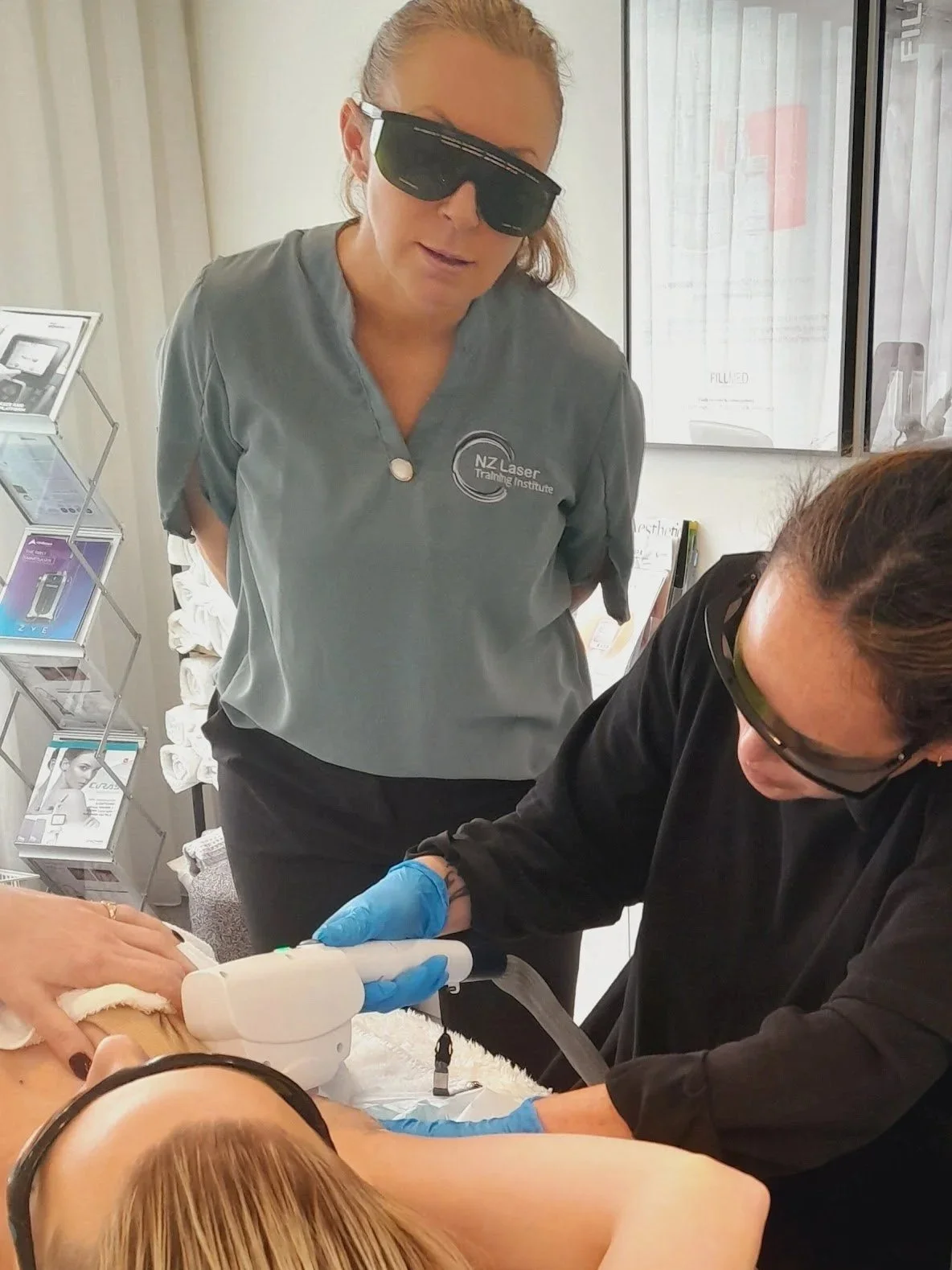 A woman receiving a laser treatment on her abdomen by a technician in a clinic. The technician is wearing black protective goggles, black gloves, and a black uniform, while the woman is lying down with black goggles on, with her head turned to the side.