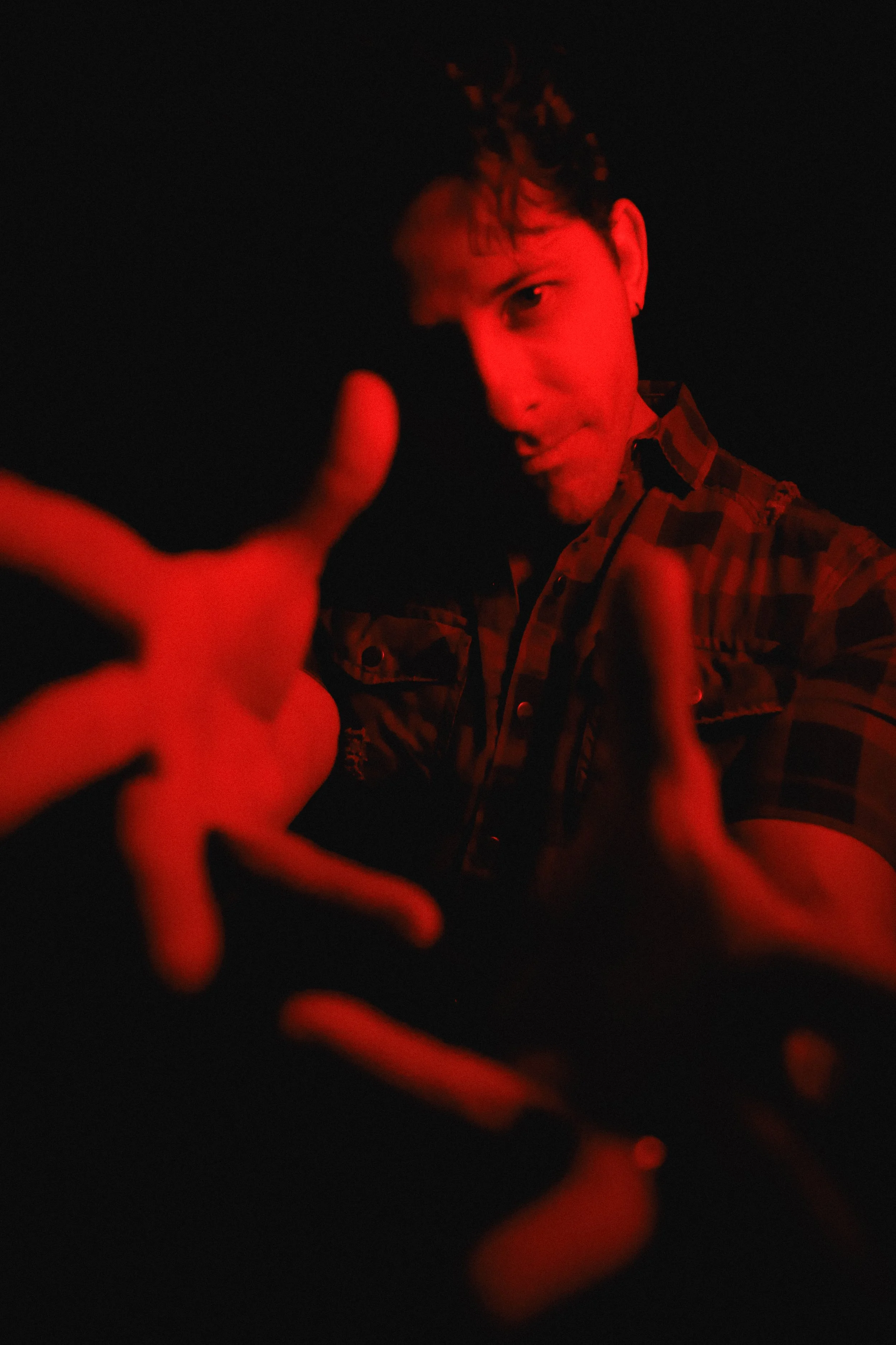 A person in a checkered shirt is reaching toward the camera, with red lighting illuminating their face and hands, against a dark background.