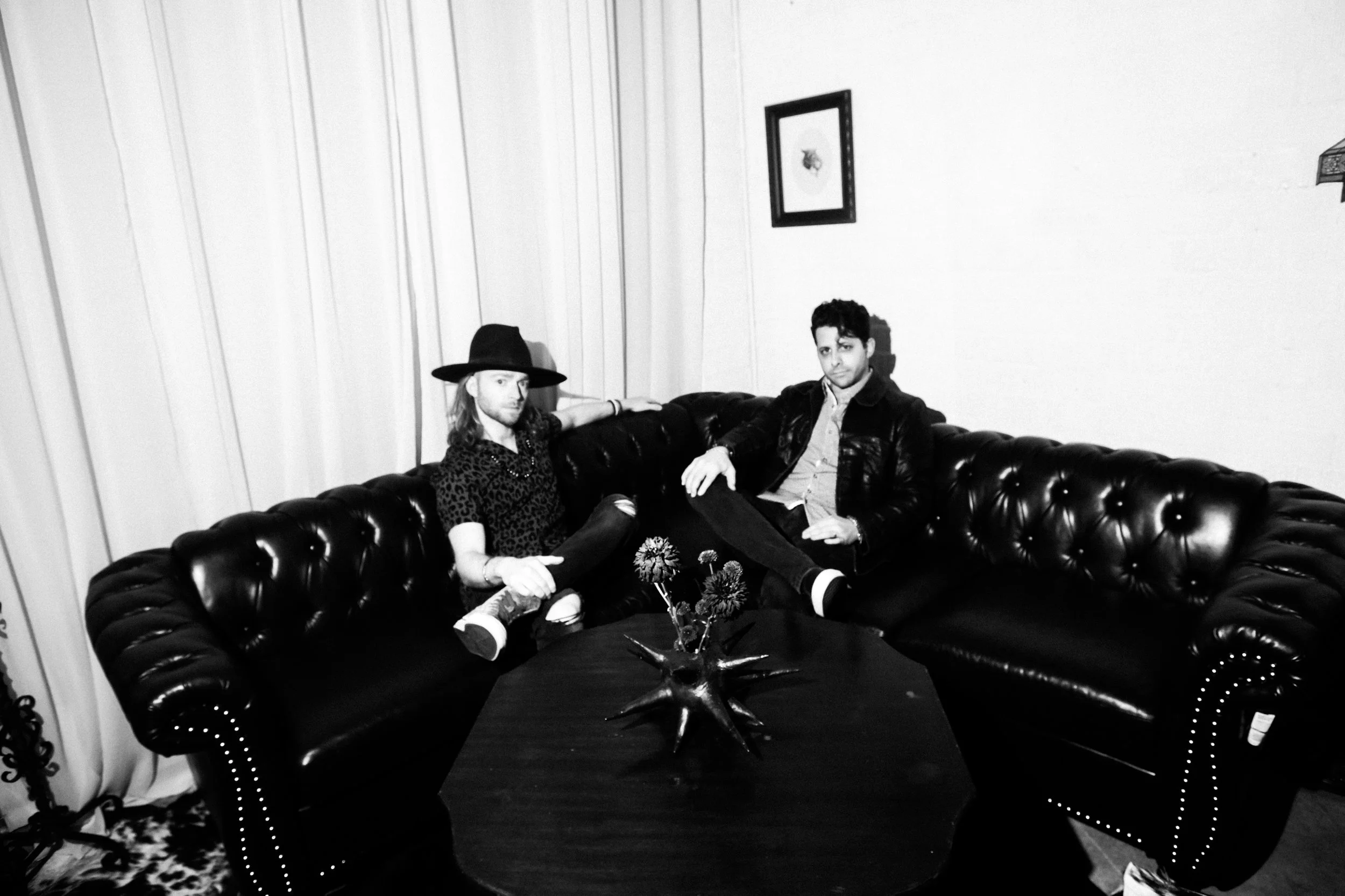 Two men sitting on a black leather couch in a room with white curtains and a framed picture on the wall. One man has long hair and is wearing a hat and patterned shirt, while the other man has short hair and is wearing a jacket and collared shirt. There is a decorative star-shaped object on the table in front of them.