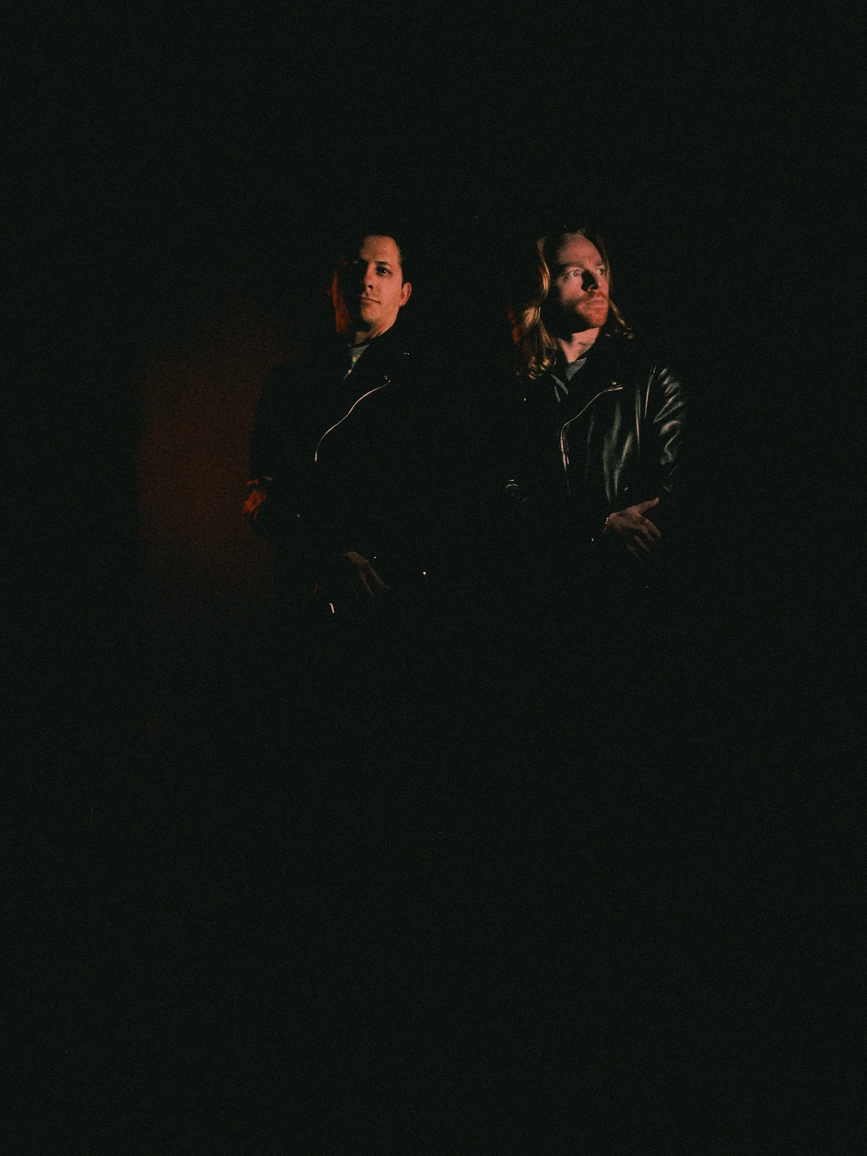 Two men standing in dim lighting, both wearing leather jackets, with serious expressions.
