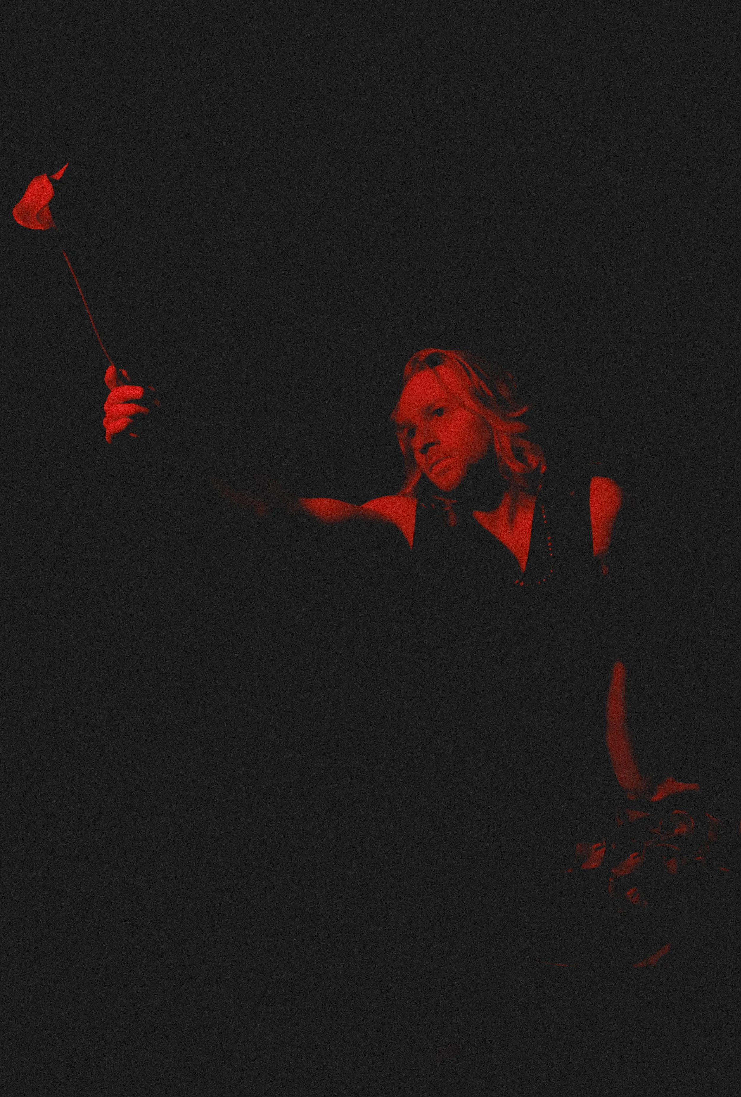 A person with long hair appears to be performing on stage, with a dark background and red lighting, holding a small red object or prop in their hand.