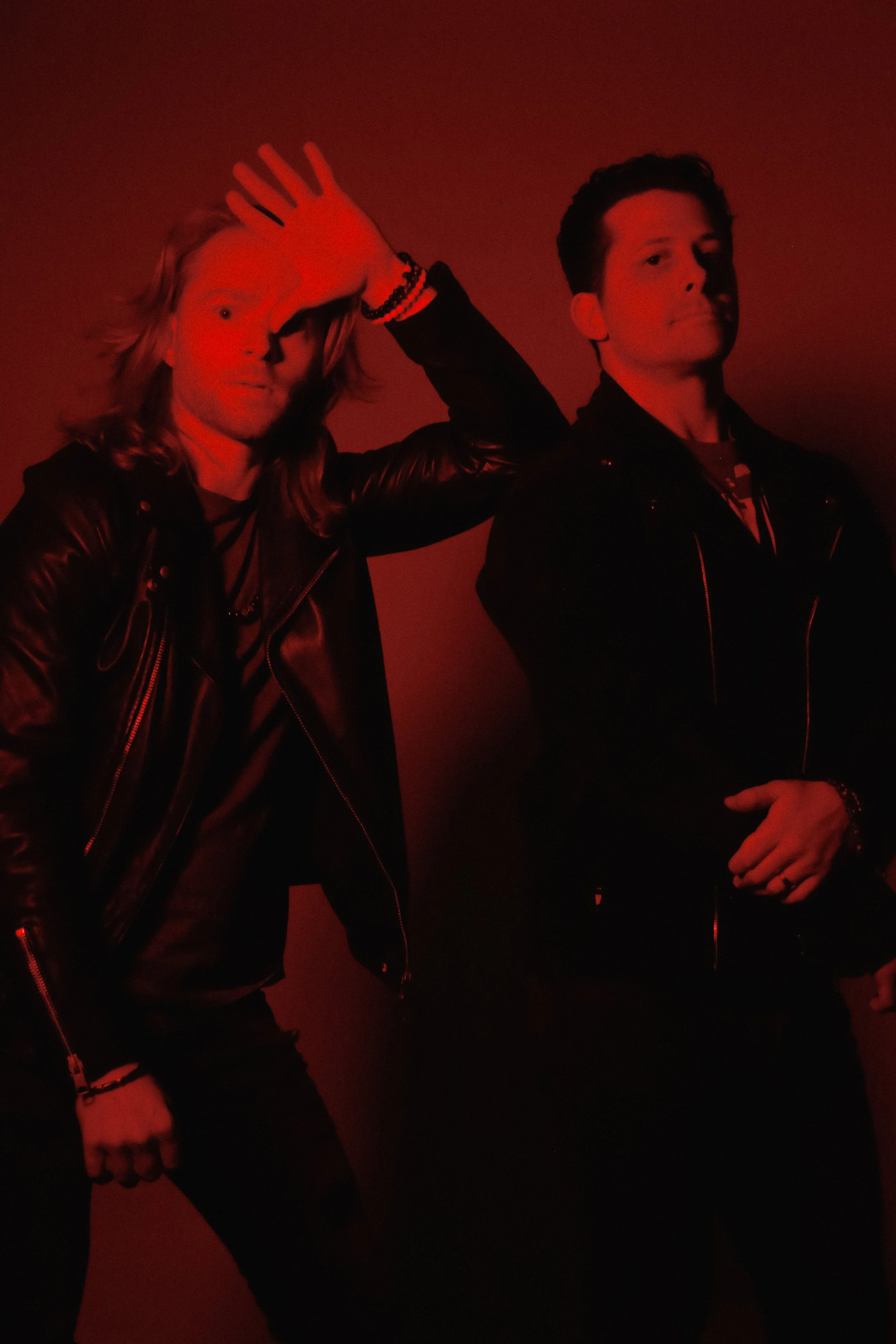Two men standing against a red background, one with long hair and the other with short hair, both wearing black leather jackets.