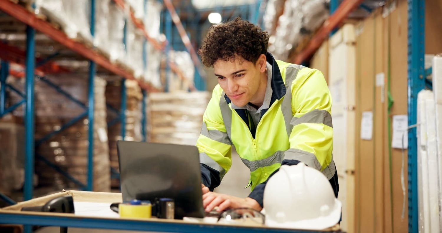 A young man in a yellow safety jacket working on a laptop in a warehouse, next to a white safety helmet on a workbench.