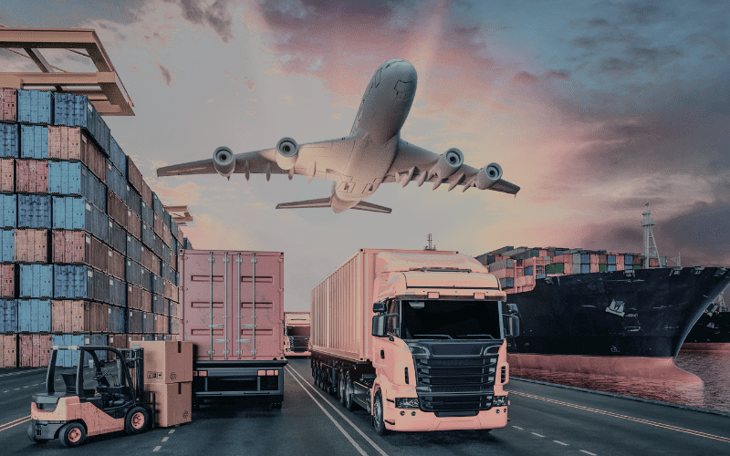 Airplane flying low over a port with shipping containers, trucks, a forklift, and a cargo ship at sunset.