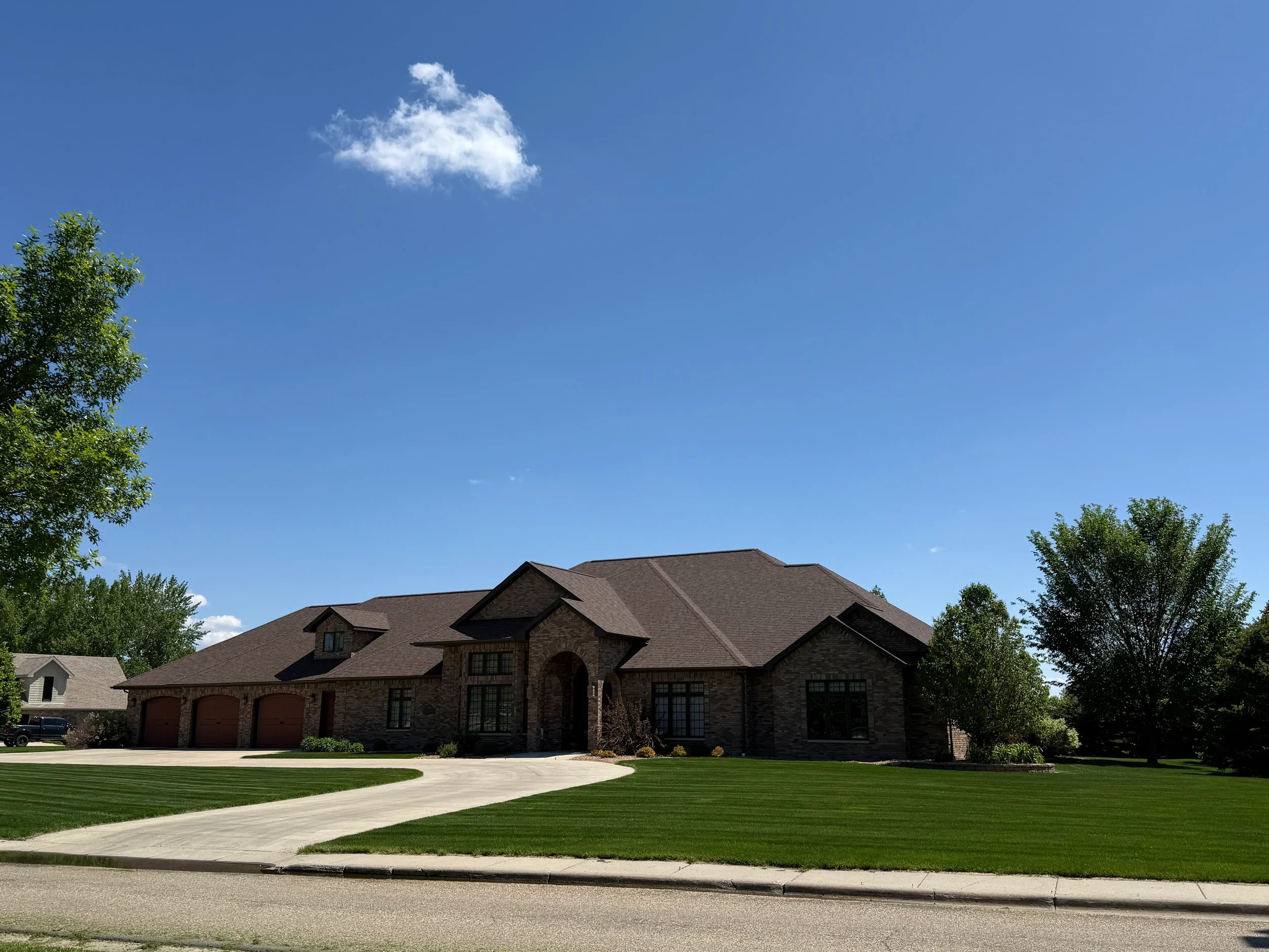 Large brick house with a three-car garage, a curved driveway, well-maintained lawn, and several trees under a clear blue sky with a few clouds.