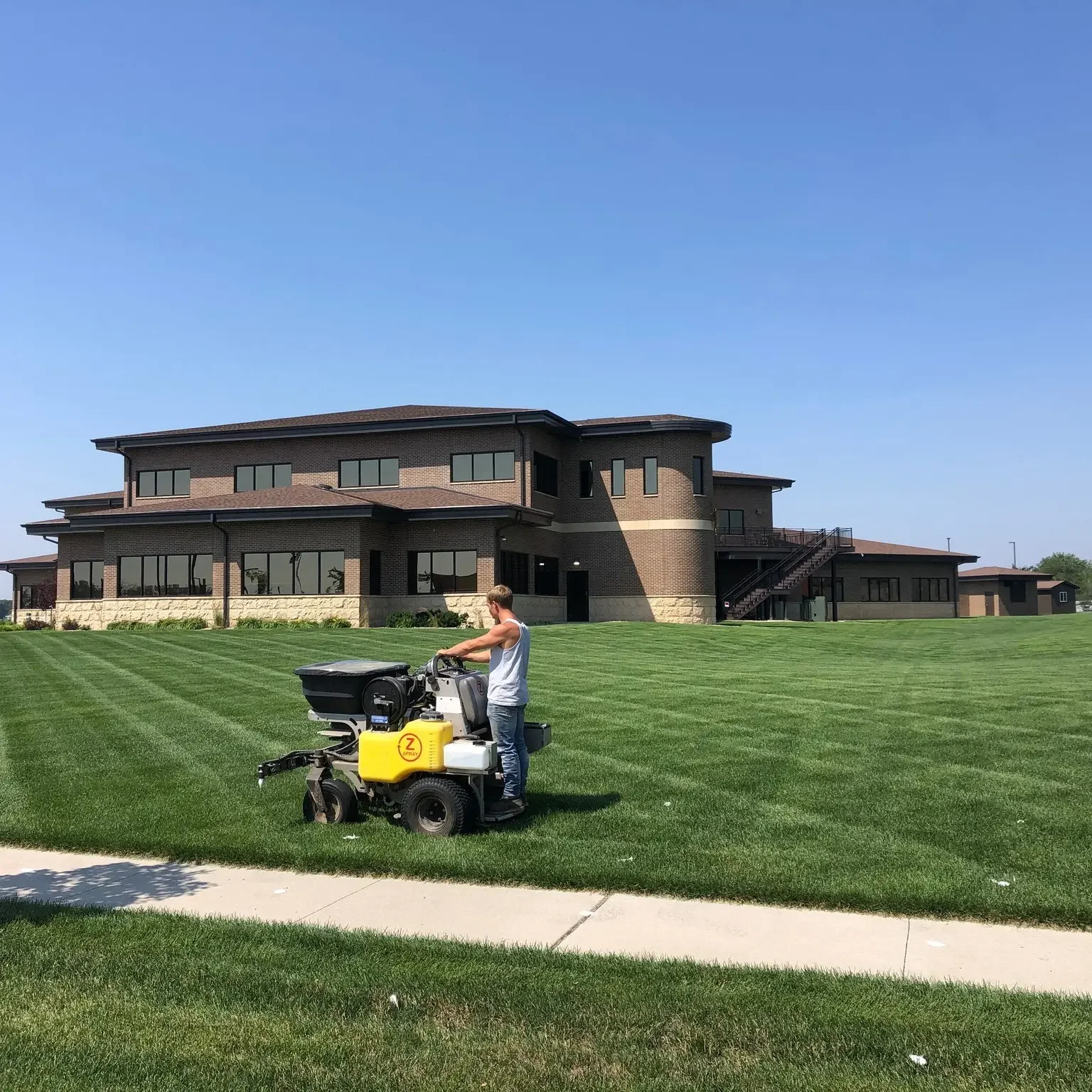 A person operating a grass mowing machine on a well-maintained lawn in front of a large modern house with multiple windows and a staircase on the side under a clear blue sky.