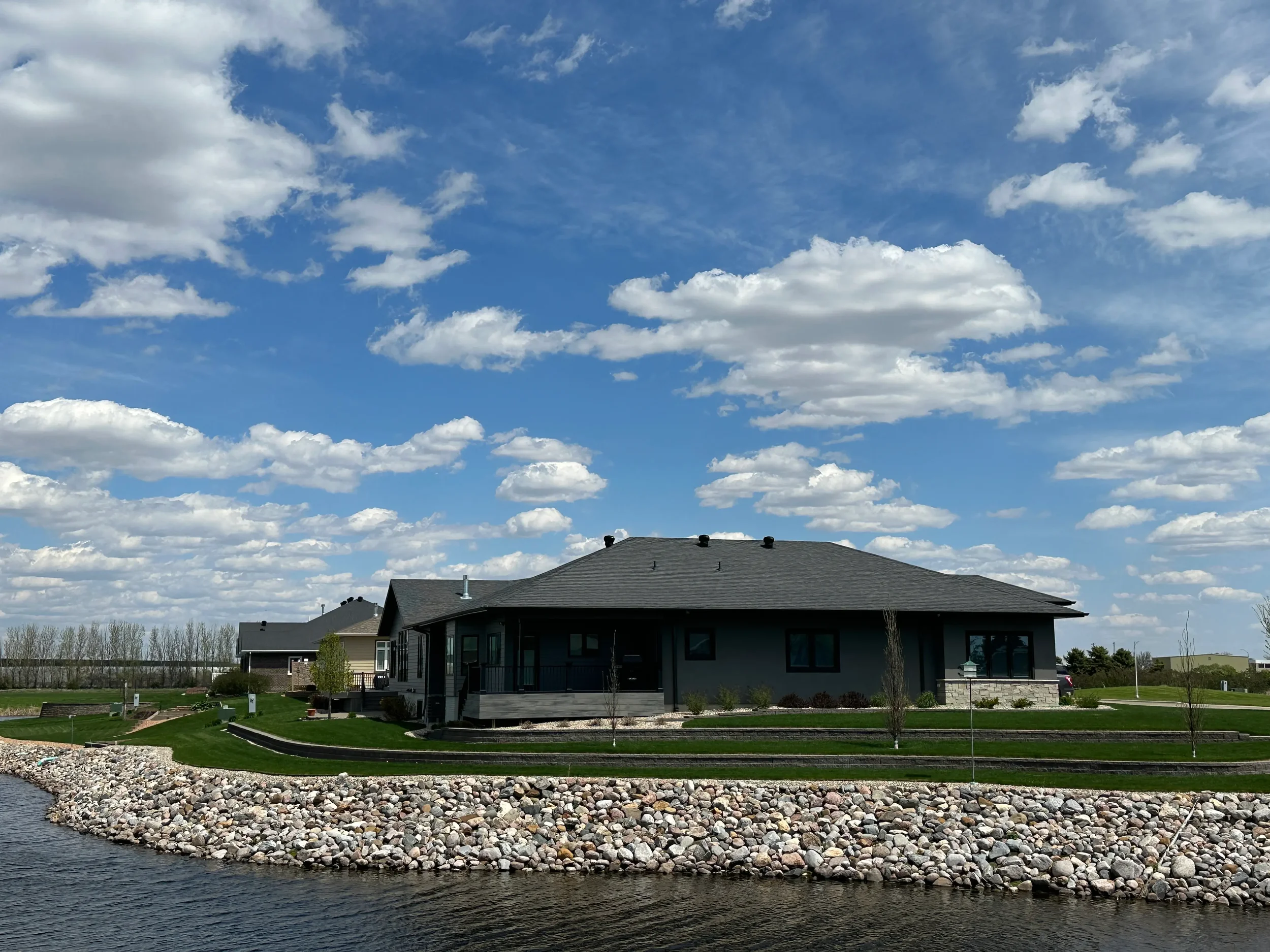 Modern house overlooking a river, surrounded by a landscaped yard with grass and young trees, under a partly cloudy sky.