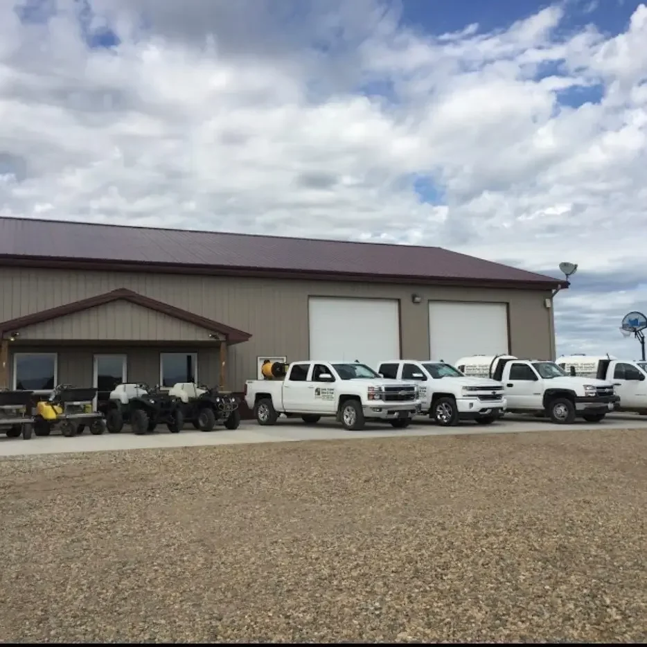 Line of white work trucks and black off-road vehicles parked in front of a large industrial building with a metal roof, under a partly cloudy sky.