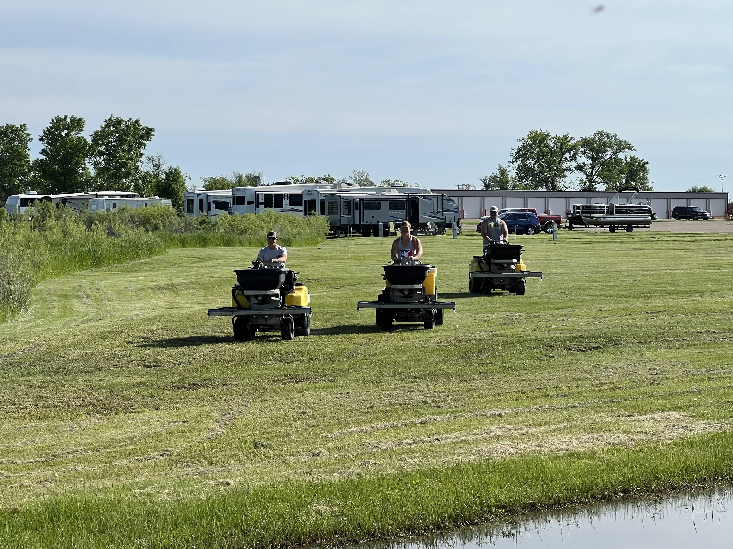 Three people riding lawn mowers across a grassy field near a pond with RVs and vehicles in the background.