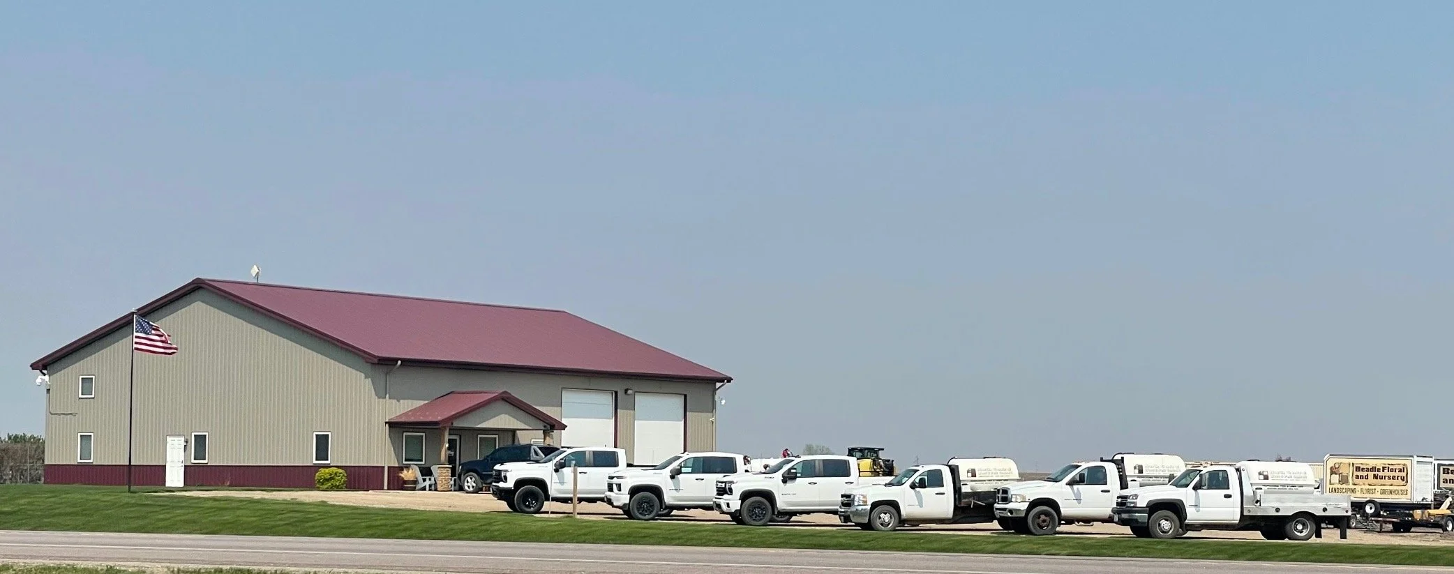 A large beige building with a red roof, American flags, several white pickup trucks, and utility vehicles parked in front. The sign on the right indicates it is a floral and nursery business.