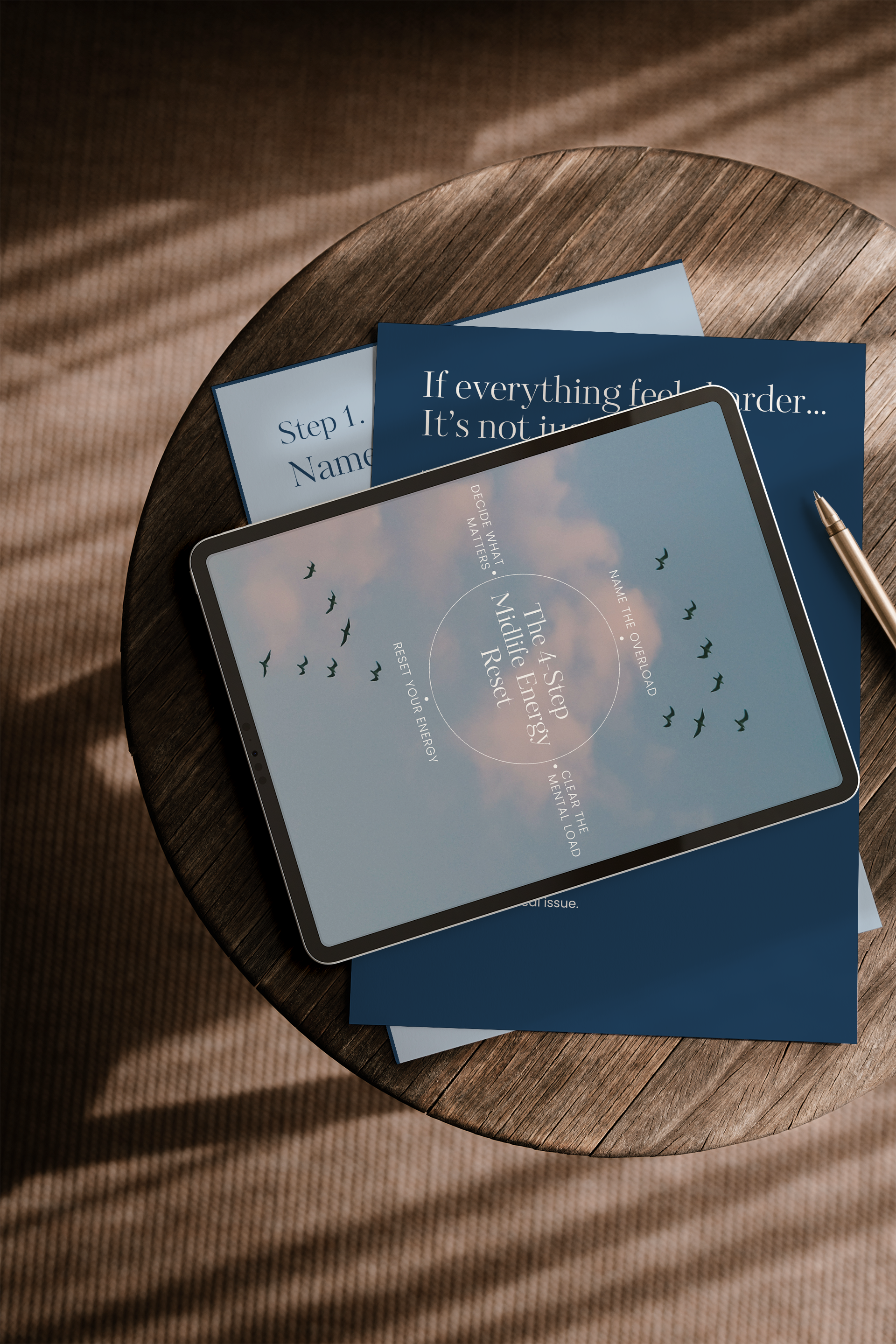 A tablet displaying a mindfulness and meditation app titled 'The 4 Step Mindful Energy Reset' is on top of blue informational sheets on a wooden table. The app includes steps like 'Decide What Matters' and 'Clear the Mental Load.' There is a gold pen beside the tablet, and sunlight casts shadows on the table and carpet.