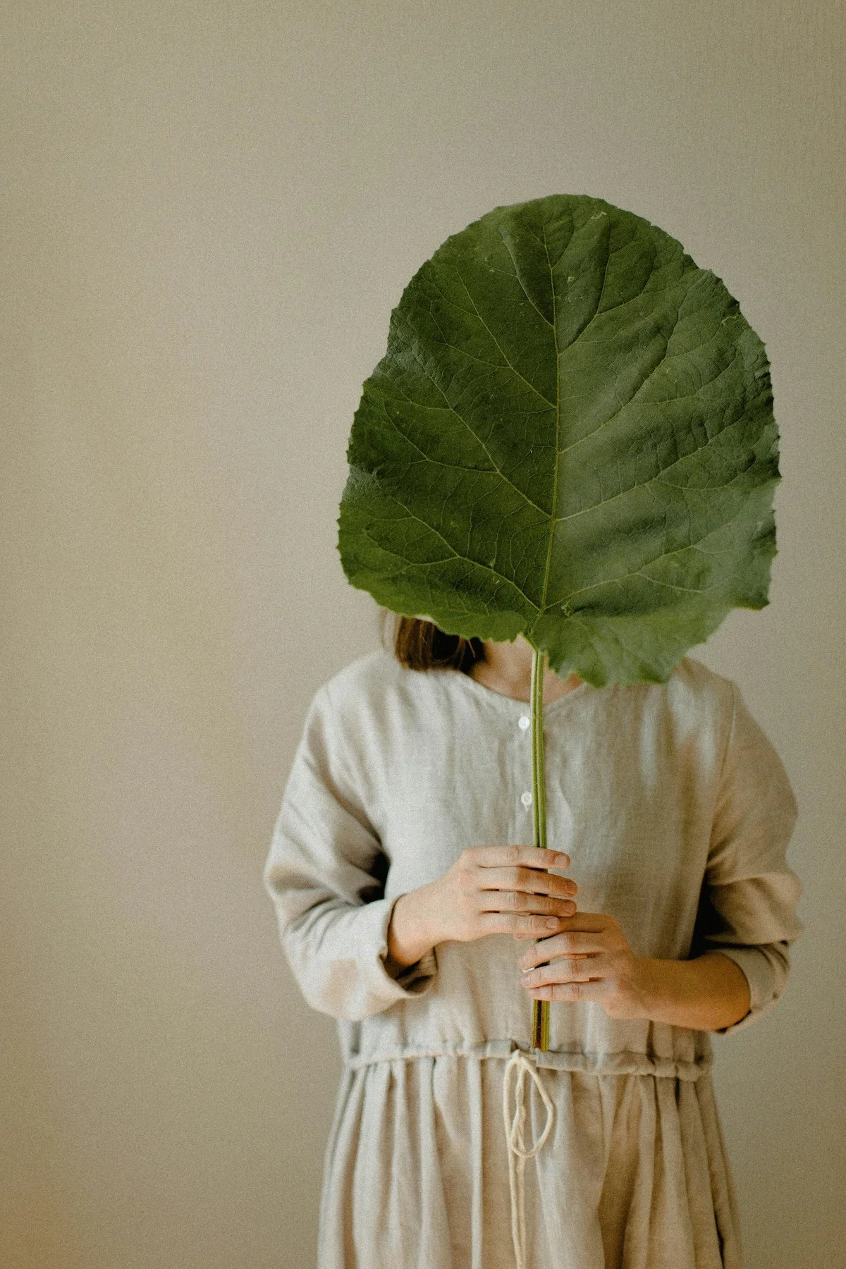 Person wearing a beige dress holding a large green leaf in front of their face, obscuring their face and head.
