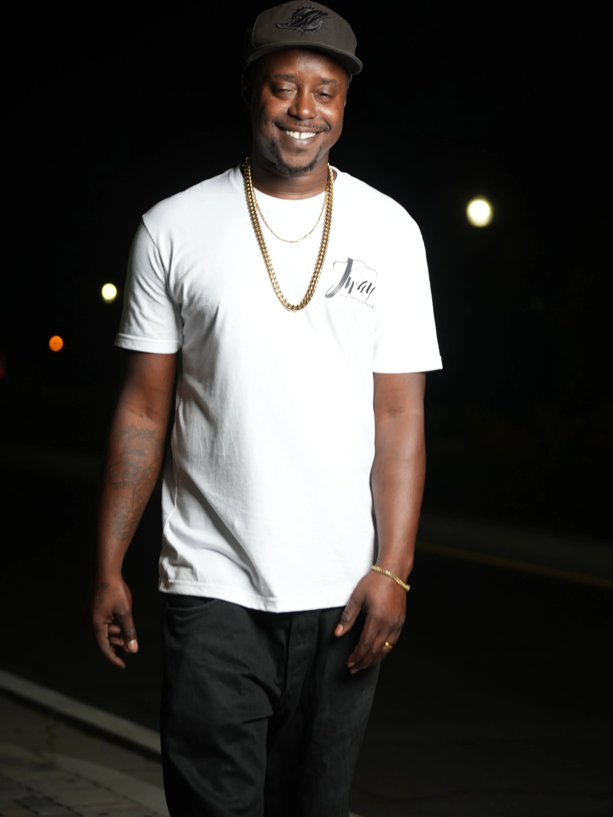 A man standing outdoors at night, wearing a white T-shirt, black pants, a gray cap, gold chains, and a gold bracelet, smiling at the camera.
