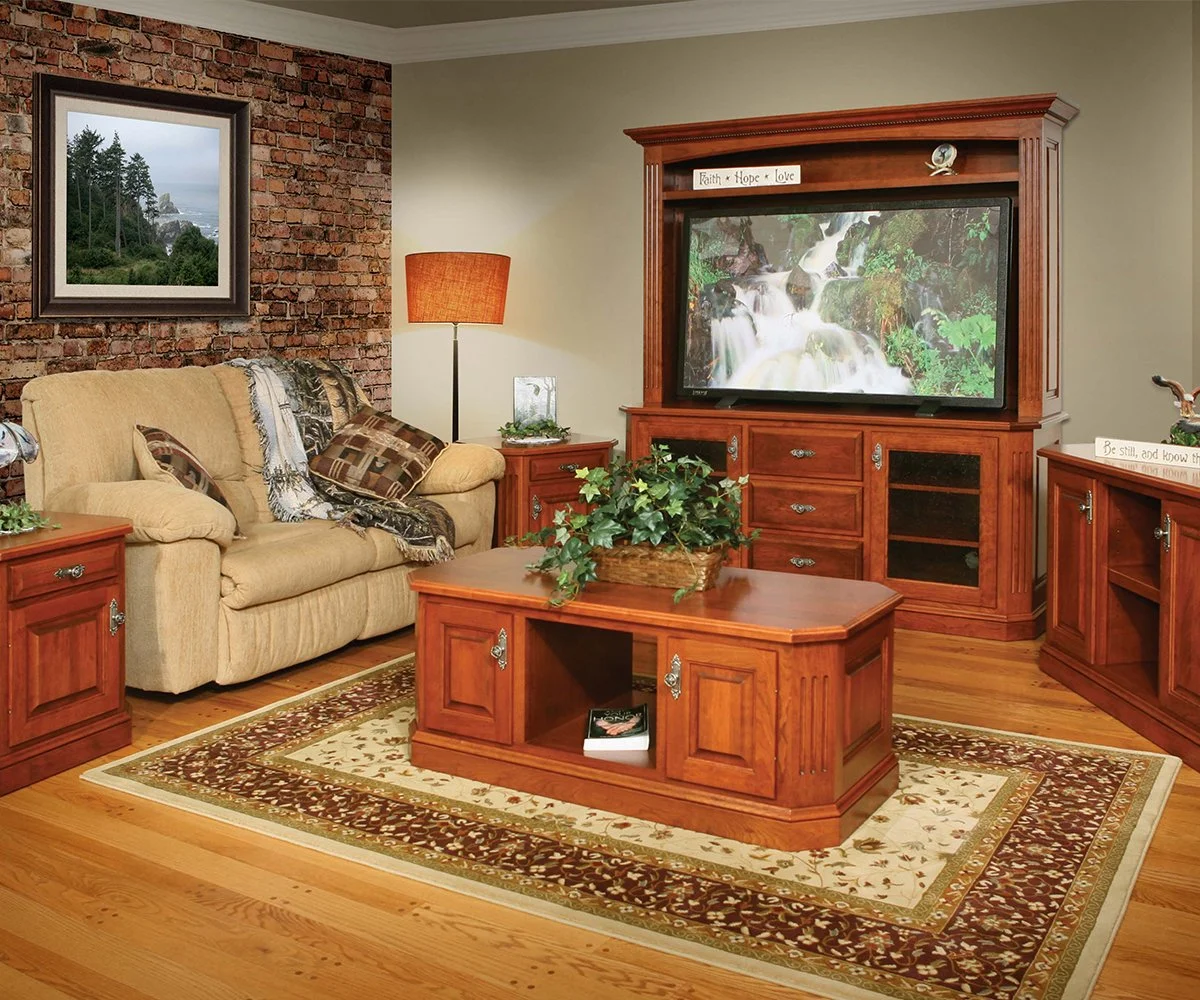 Living room with brick accent wall, beige sofa with pillows, wooden coffee table with plants, wooden TV stand with a large flat-screen TV displaying a waterfall scene, framed landscape picture on the brick wall, and a tall orange lampshade.