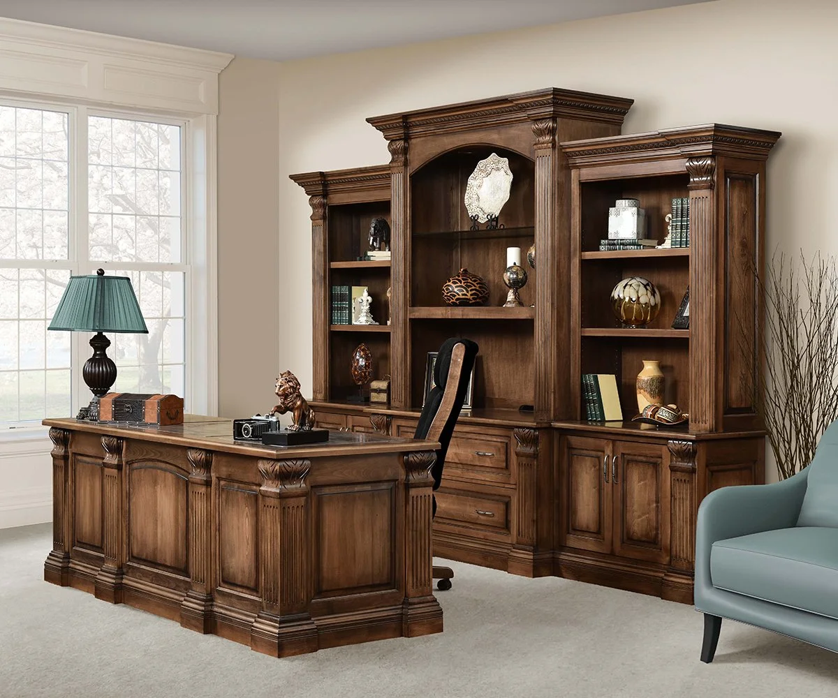 A traditional home office with a wooden desk, a black office chair, and a large wooden bookshelf filled with decorative items and books. There is a window with white trim, a teal lamp on the desk, and a light blue armchair in the corner.
