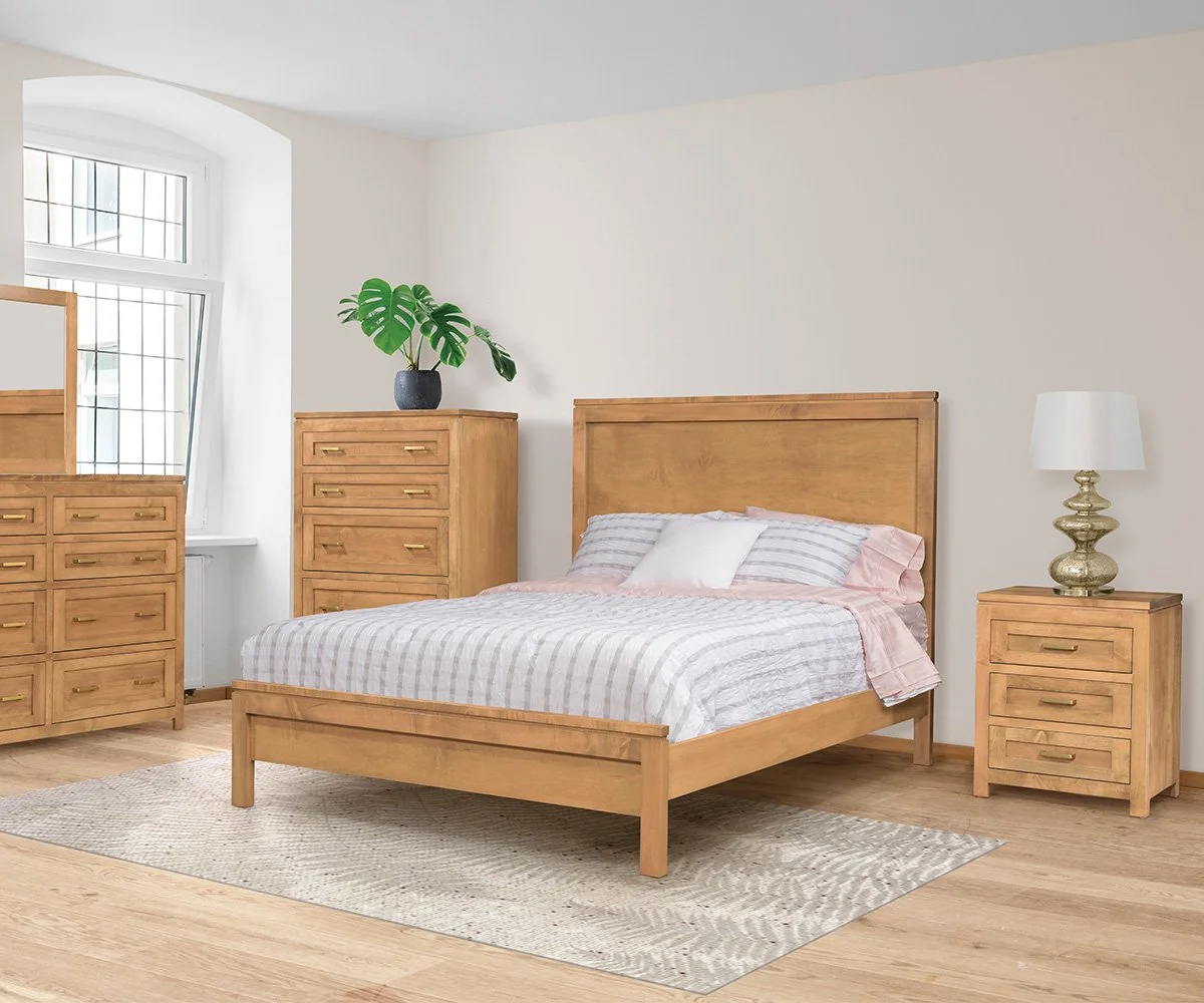 A bedroom with light wood furniture including a bed, a nightstand, a tall dresser, and a shorter dresser with a mirror. The bed has striped bedding and pillows. There is a white lamp on the nightstand and a green plant on the tall dresser. The room has a large window with a semi-angled windowpane design, white walls, and a hardwood floor with a beige rug.