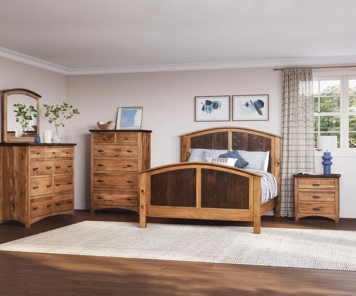 A bedroom with wooden furniture, including a bed, dresser, tall chest, and nightstand. The bed has striped linens and decorative pillows. There are framed abstract artworks above the bed. A window with curtains lets in natural light, and a blue vase and plant decor are on the furniture. A light-colored area rug covers part of the wooden floor.