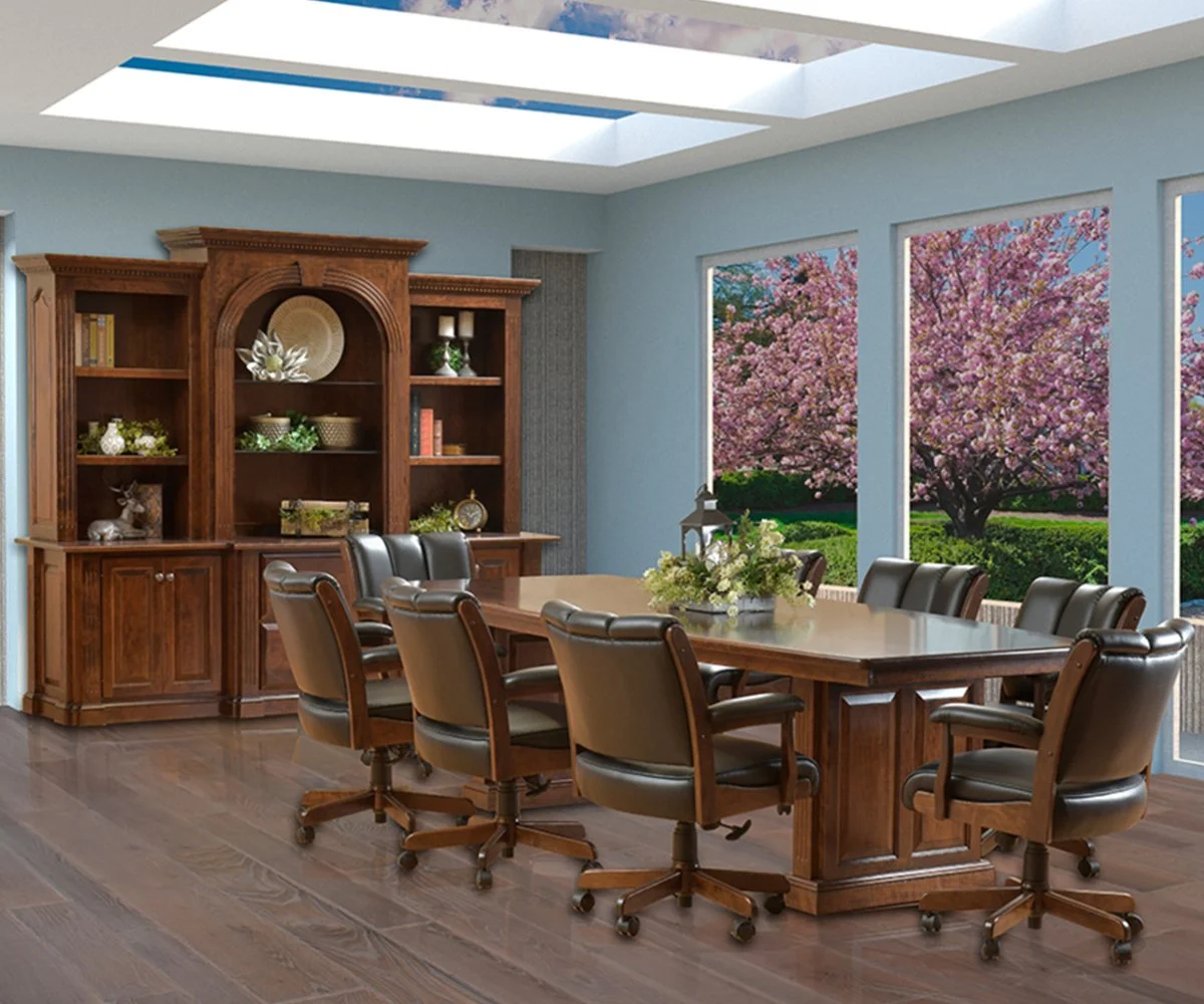 A conference room with a large wooden table surrounded by nine black office chairs, a wooden bookshelf against a blue wall, and three large windows showing blooming pink cherry blossom trees outside.