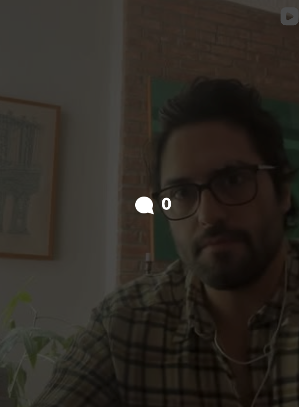 A man with dark hair, glasses, and a beard wearing headphones and a plaid shirt indoors, with a brick wall and artwork behind him.