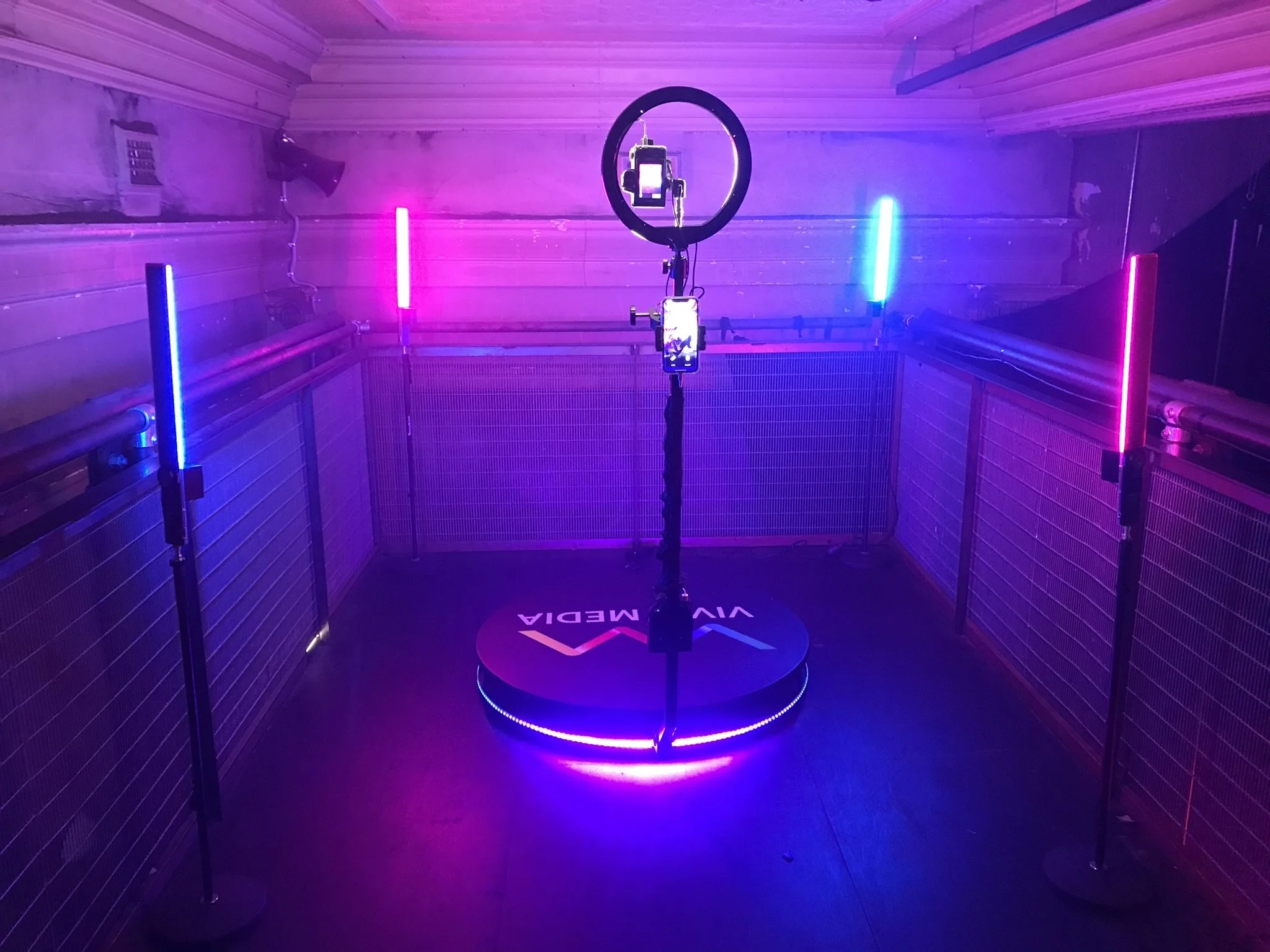 Virtual reality gaming setup with a central ring light, a smartphone mounted in the middle, and colorful vertical LED lights in a room with ceiling pipes, labeled 'VIVA MEDIA' on the platform