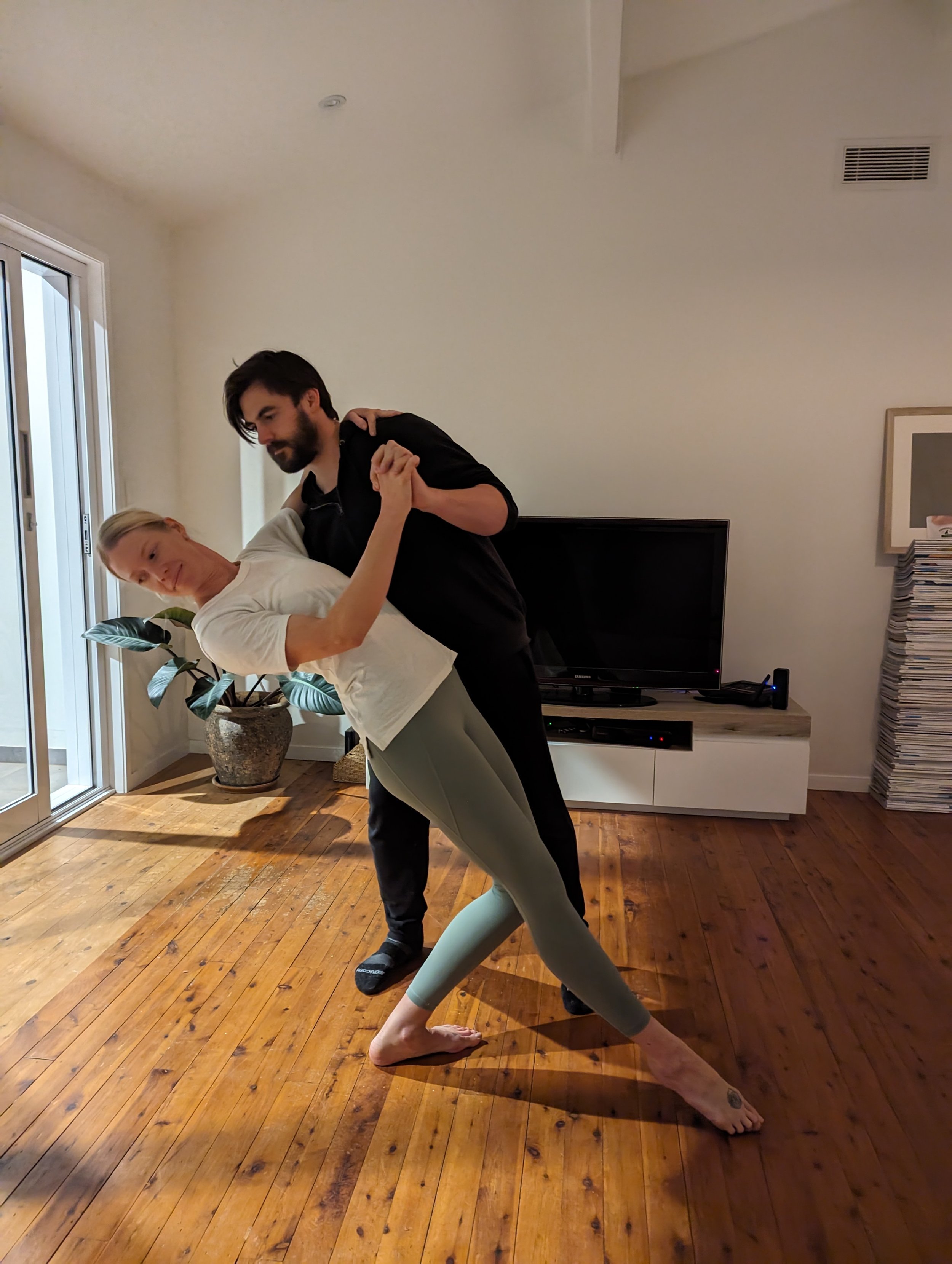 A woman and man dancing in a living room. The woman is leaning back with her left foot forward and right foot behind, while the man holds her, supporting her with his left arm around her back.