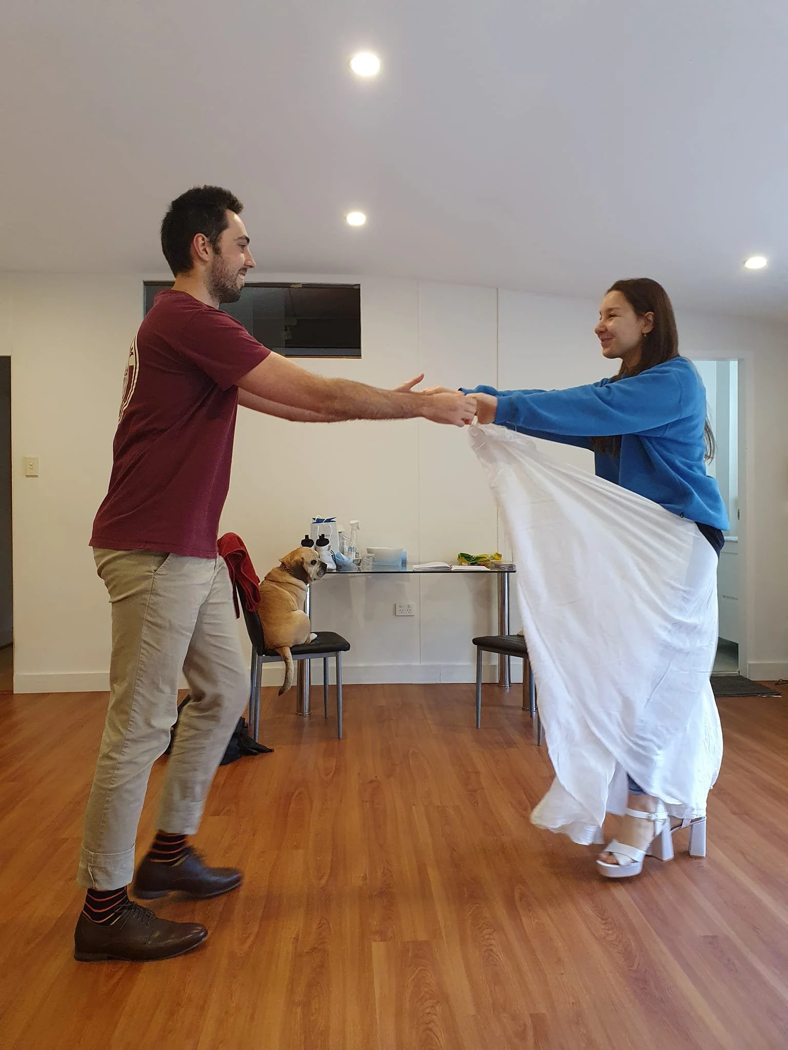 A man and woman are dancing together indoors, holding hands and smiling. A dog is sitting on a chair in the background.