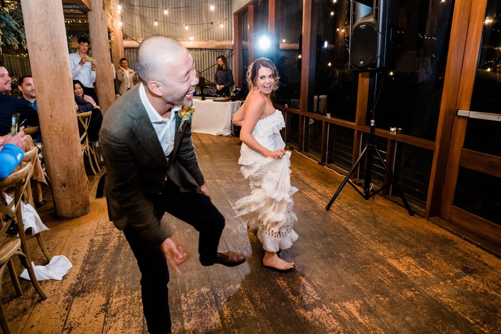 At a wedding reception, a man in a suit and a woman in a white dress are dancing together, smiling and enjoying themselves. Other guests are sitting and standing around, watching and taking photos of the dance. The venue has wooden floors and walls, 
