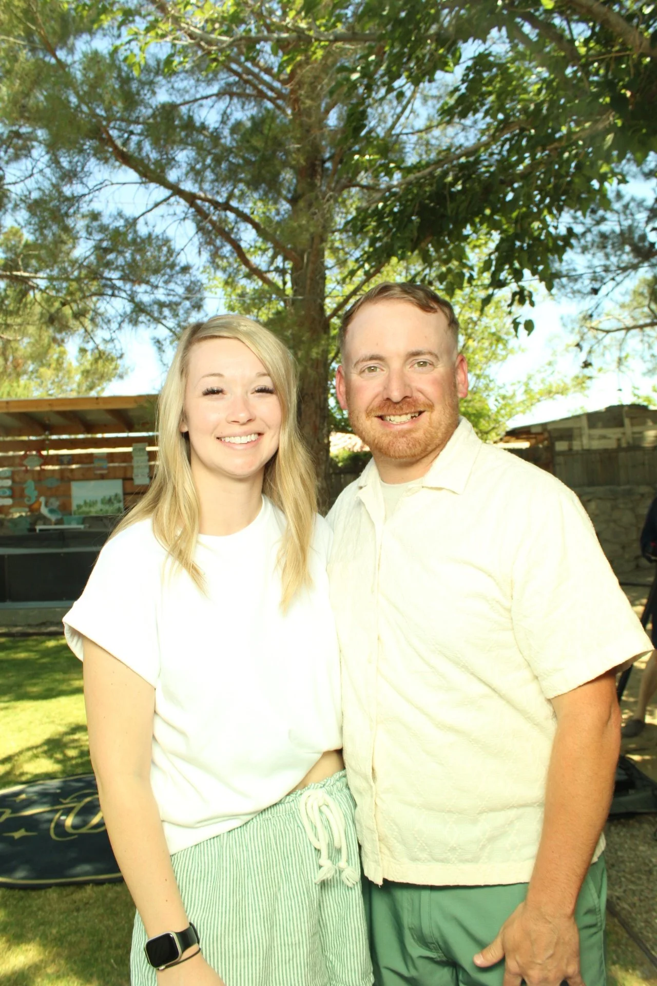 A smiling young woman with long blonde hair standing next to a smiling man with short reddish hair and beard outdoors. They are standing under a tree in a backyard with a wooden fence and a covered patio in the background.