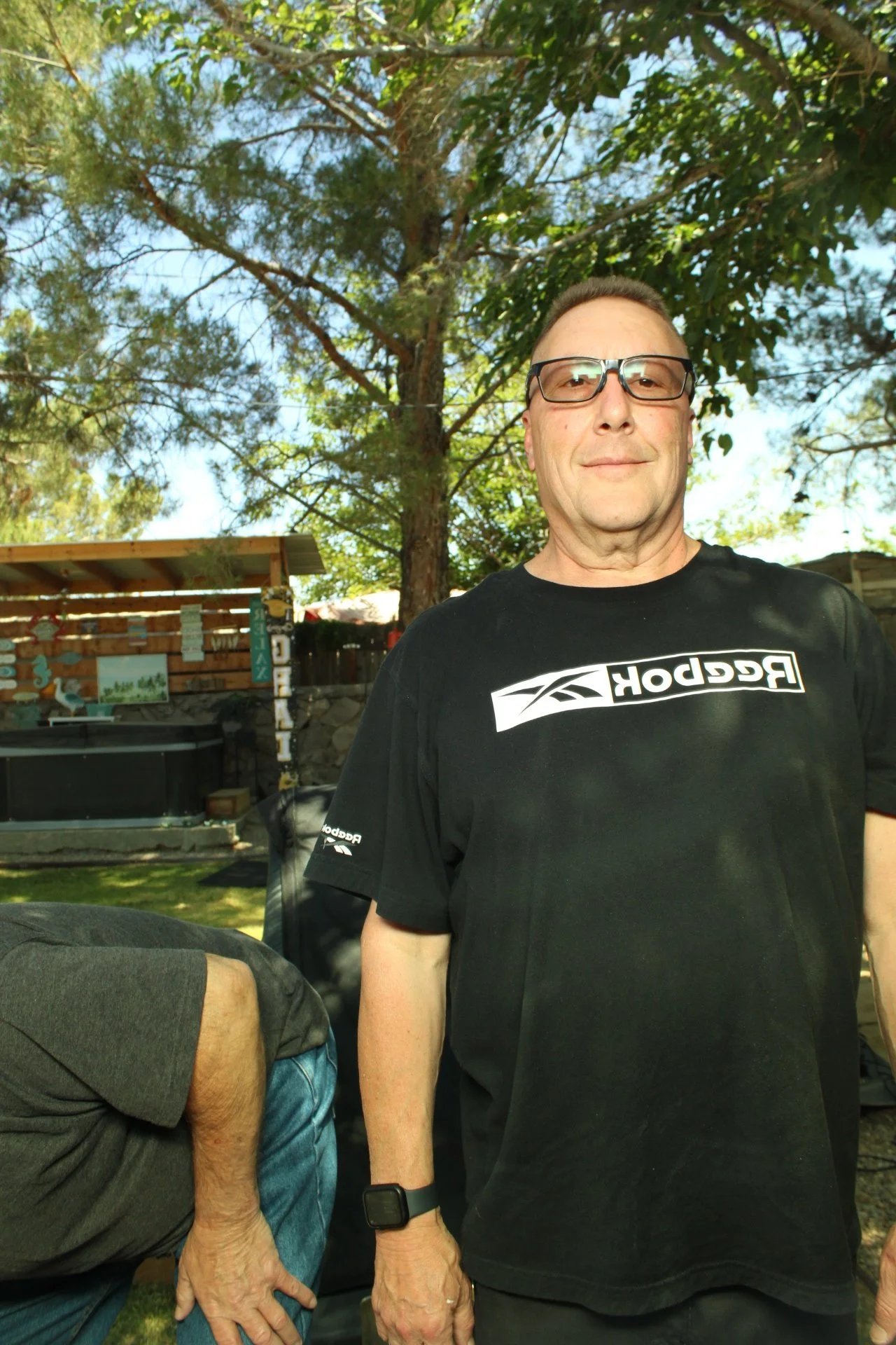 A man with glasses and a black Reebok t-shirt stands outdoors in a backyard with trees, a wooden fence, and a hot tub in the background. Another person's arm and torso are partially visible in the foreground.
