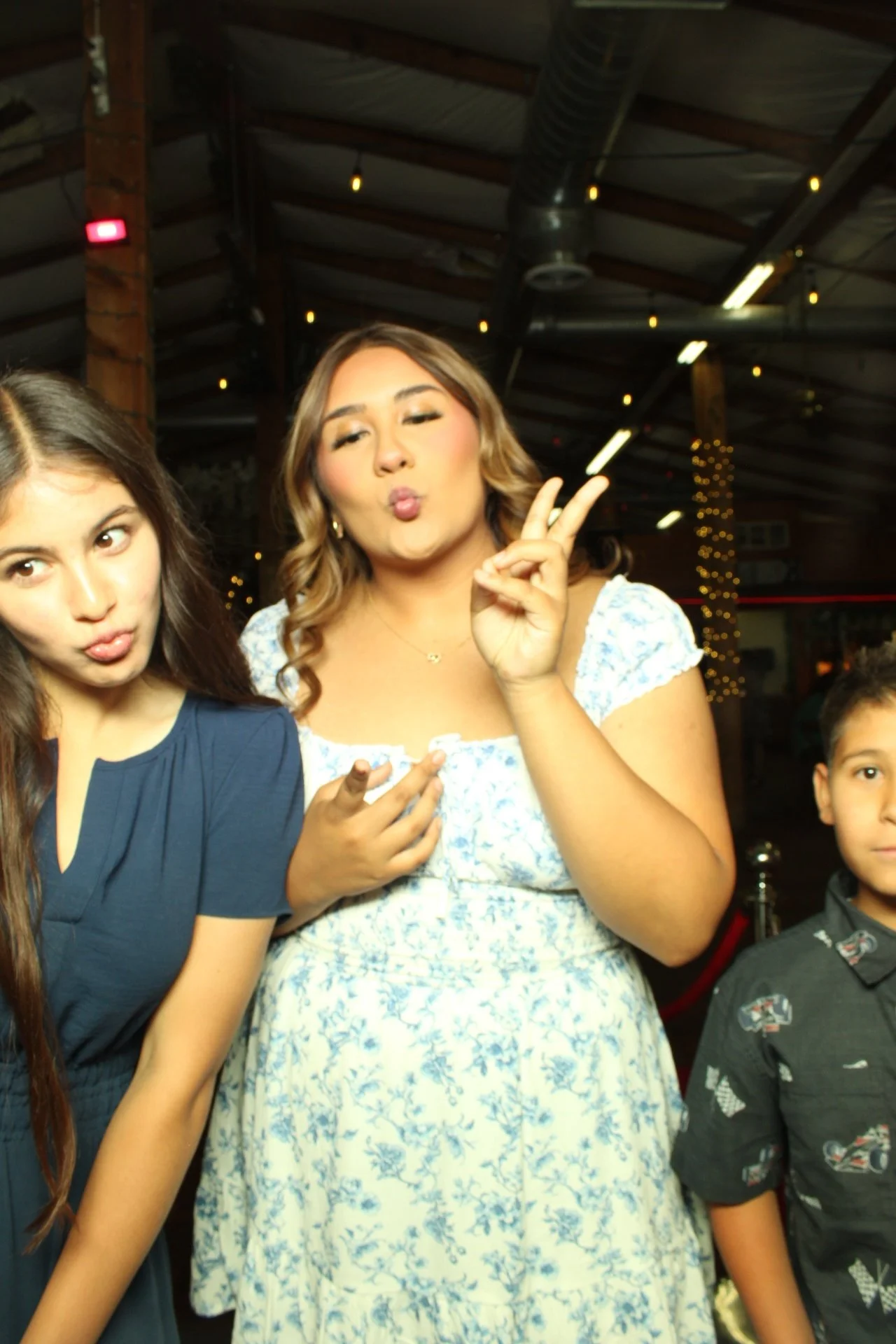 Two young women and a young boy posing at an indoor event with wooden beams and string lights in the background. The woman in the middle is making a peace sign and puckering her lips, wearing a light blue floral dress. The woman on the left has long dark brown hair, wearing a dark blue dress, and is making a similar face.