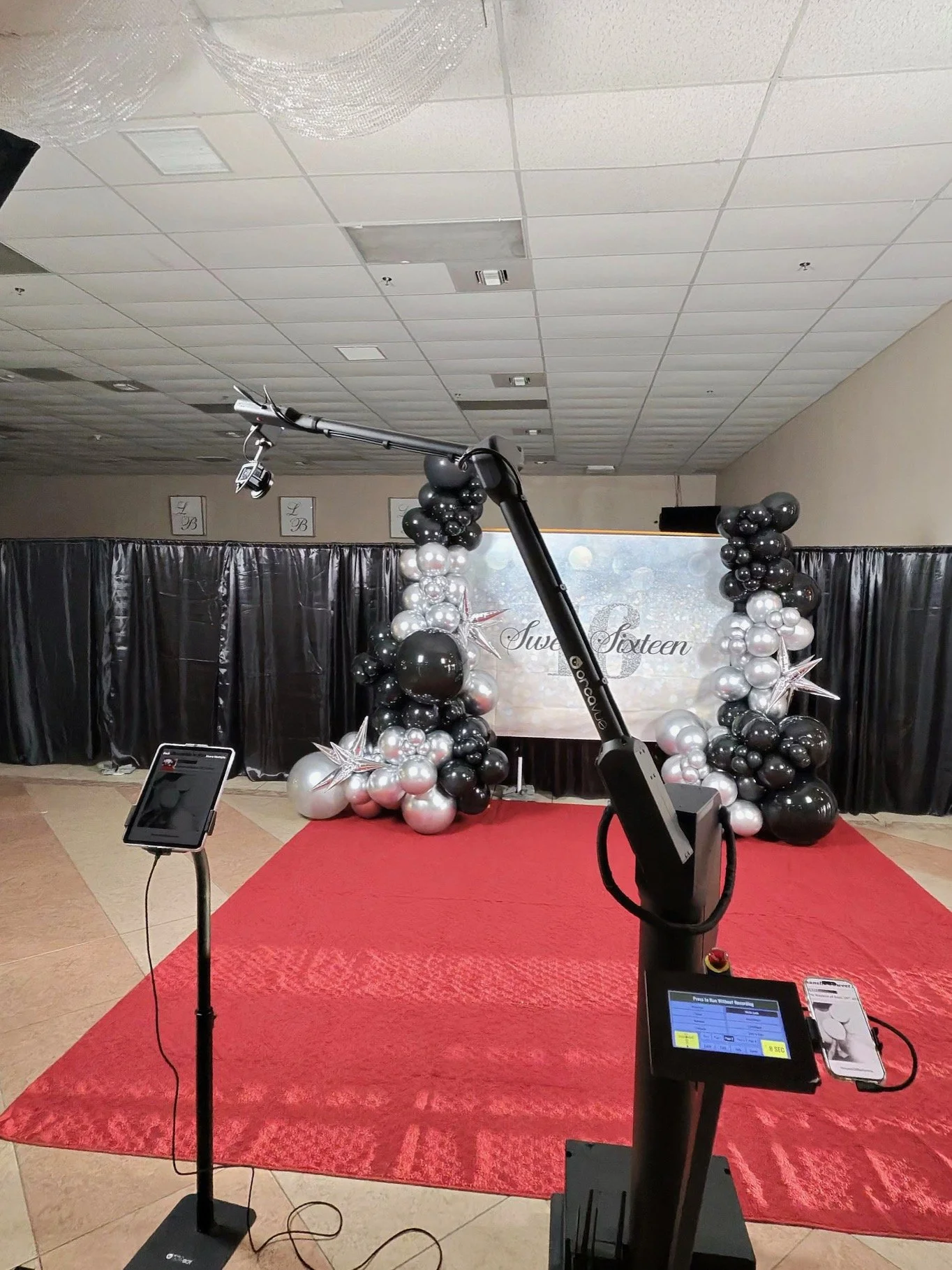 Glambot Event stage decorated with black, white, and silver balloons, with a red carpet, black curtains, and a screen in the background that reads 'Sweet Sixteen.' There are photo booth props and equipment, including a camera and lighting setup.