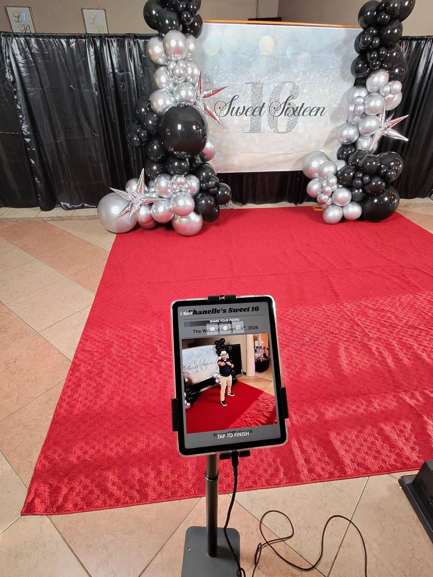 A red carpet with a backdrop featuring the words "Sweet Sixteen" and the number 16 in silver glitter, surrounded by balloon arrangements in black, silver, and white, with star-shaped decorations. Glambot Sharing station ready.