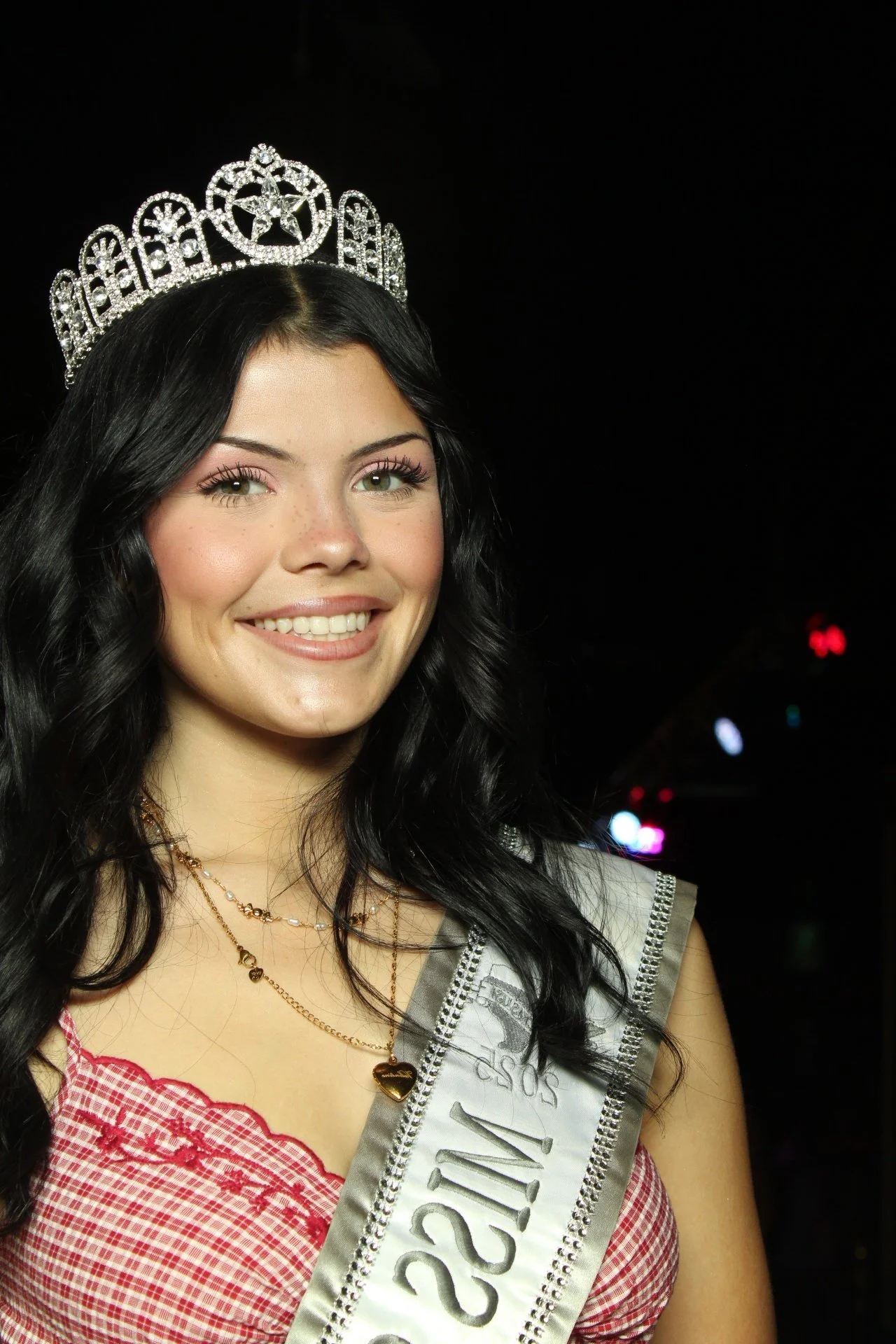 Young woman with long dark hair wearing a crown, smiling, dressed in a red gingham top, with a sash that reads 'Sofia 2023', against a dark background with colorful lights.