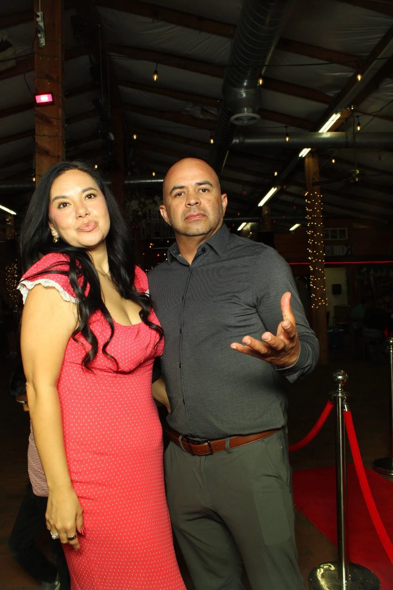 A woman in a red polka dot dress and a bald man in a gray shirt are posing at an indoor event, with string lights and wooden beams in the background.