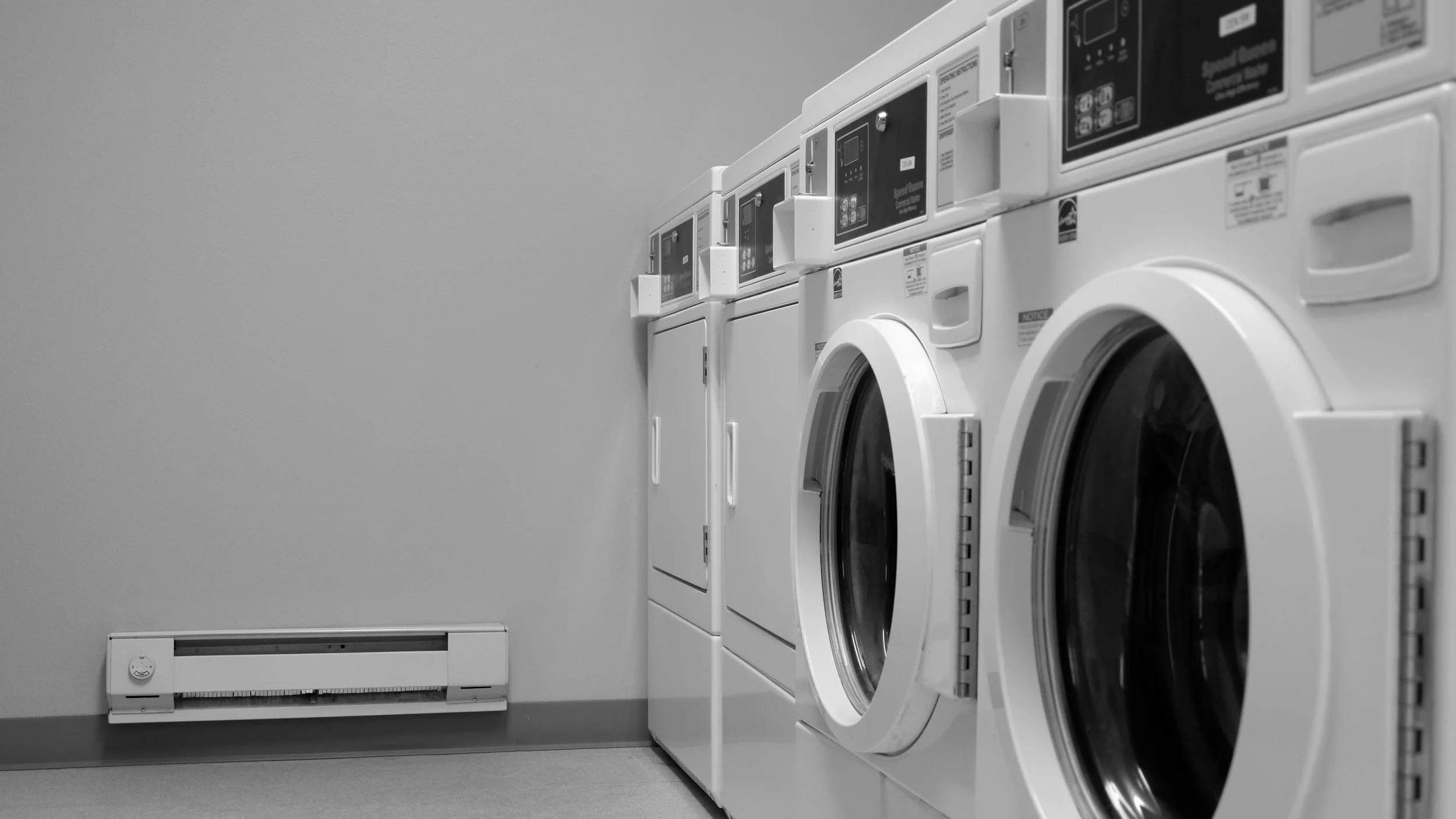 A row of washing machines lined up in a laundry room, with a plain wall and a baseboard heater visible on the left side of the image.